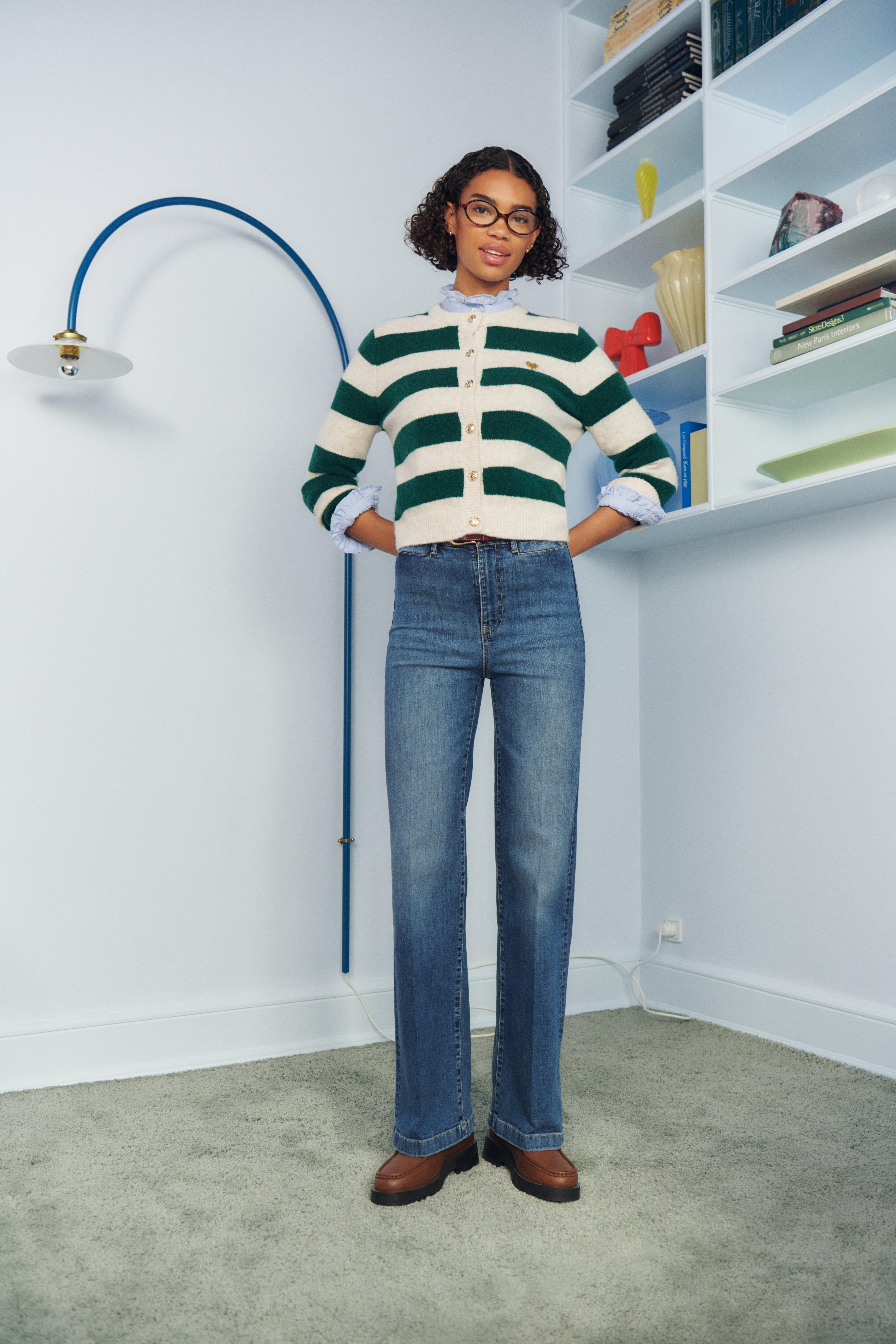 A woman standing in a corner of a room, she is wearing a striped cardigan and Lindex jeans.