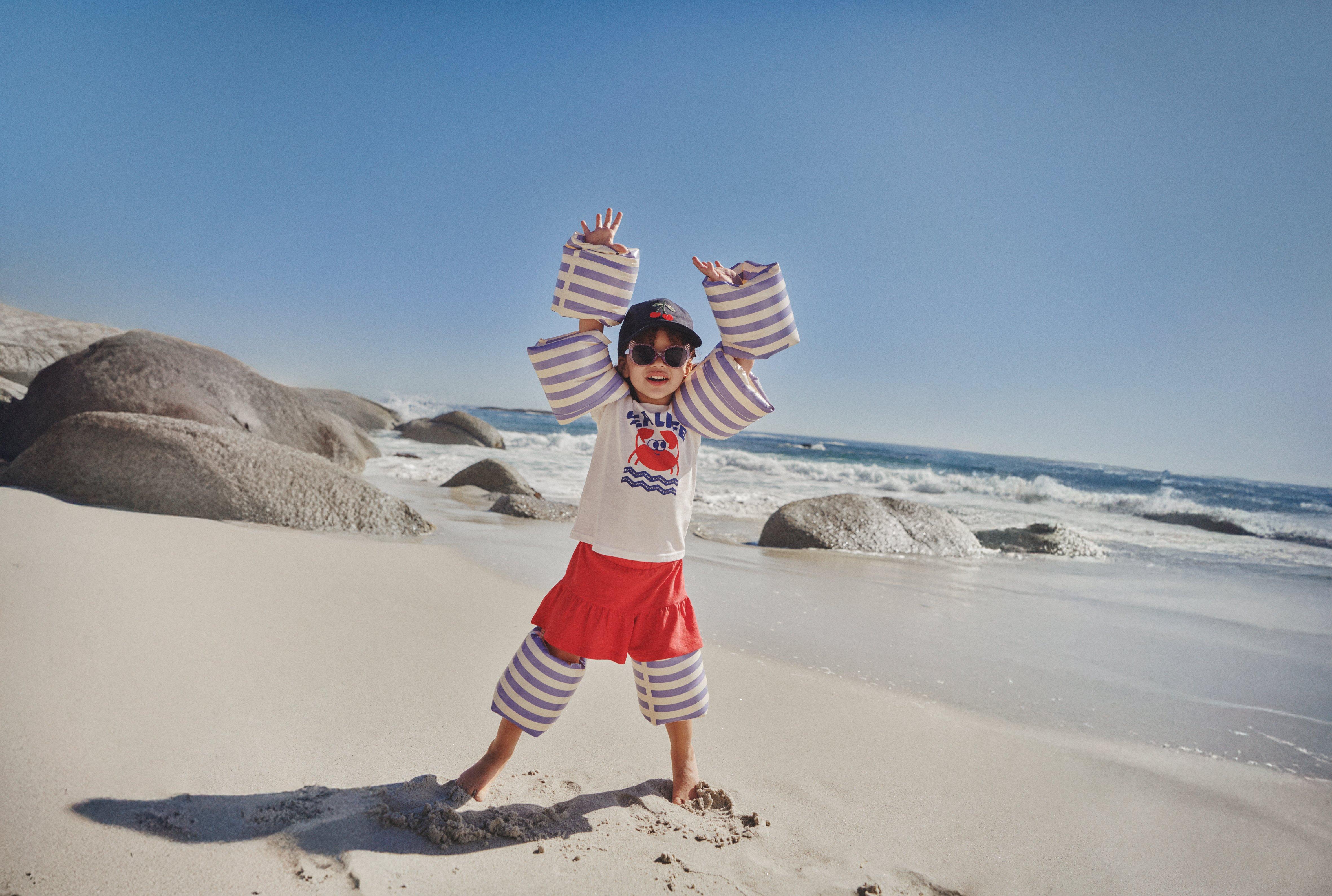 Chlapec v červených šortkách a tričku s potiskem kraba stojí na pláži. Má na rukou a nohách několik ploutví. Ruce má natažené do vzduchu.