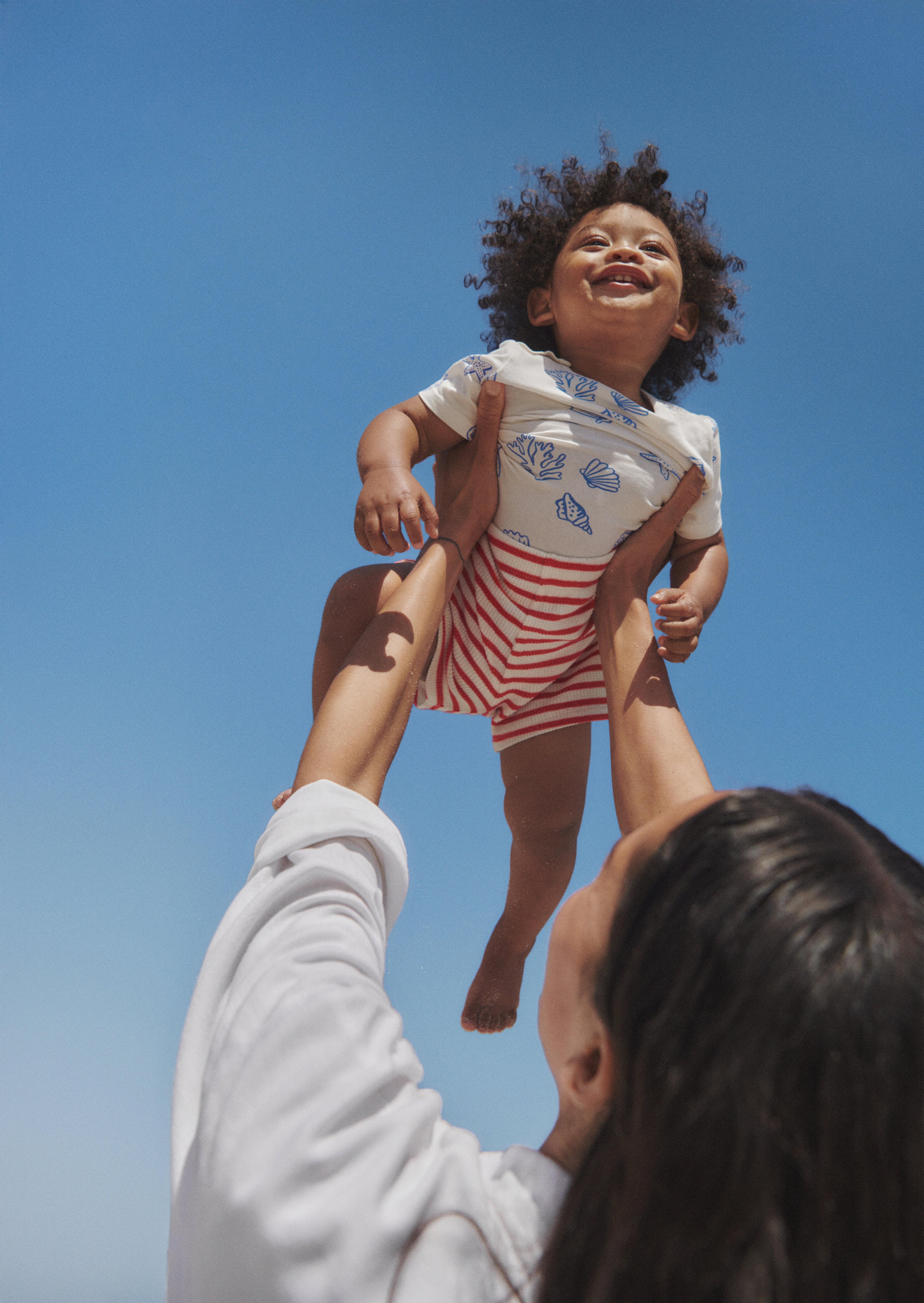  A laughing baby being held in the air by his mother. The baby is wearing a pair of red striped shorts and a t-shirt with blue shells.