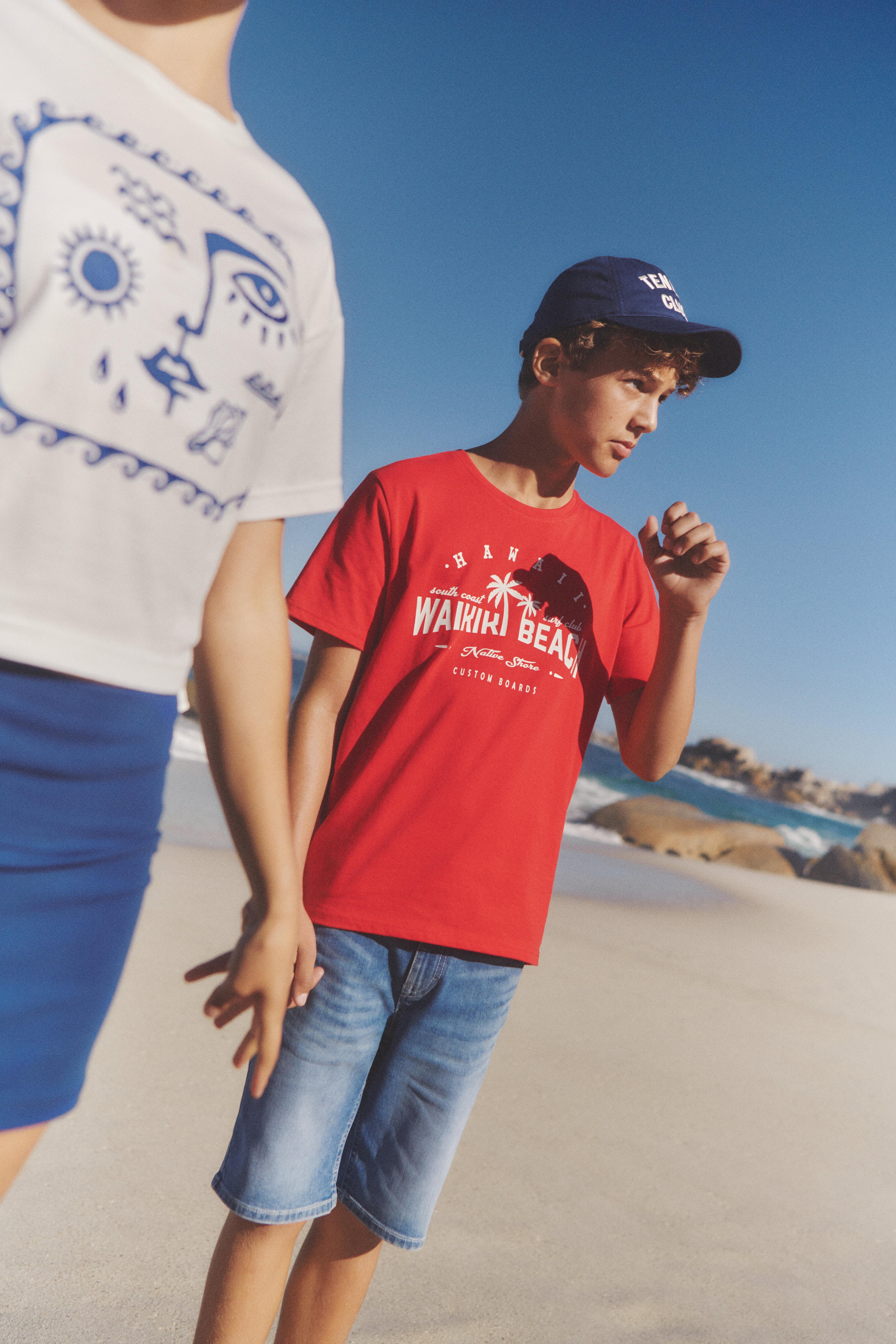 Chlapec v dlouhých džínových šortkách a červeném tričku s potiskem jde po pláži.