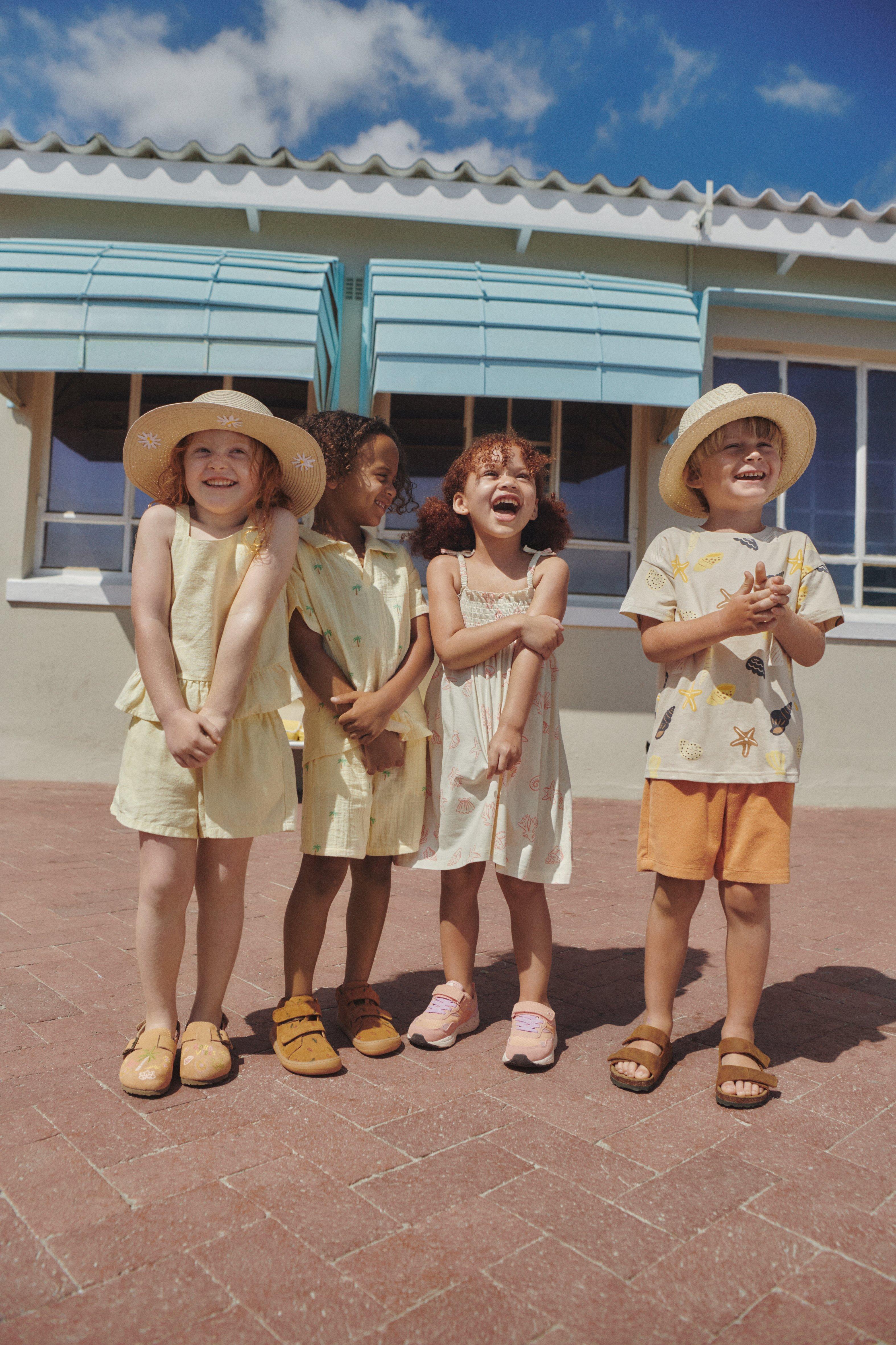Čtyři děti oblečené v letním oblečení stojí a smějí se před fasádou domu. Slunce svítí. Dvě z dětí mají na sobě sluneční klobouky.