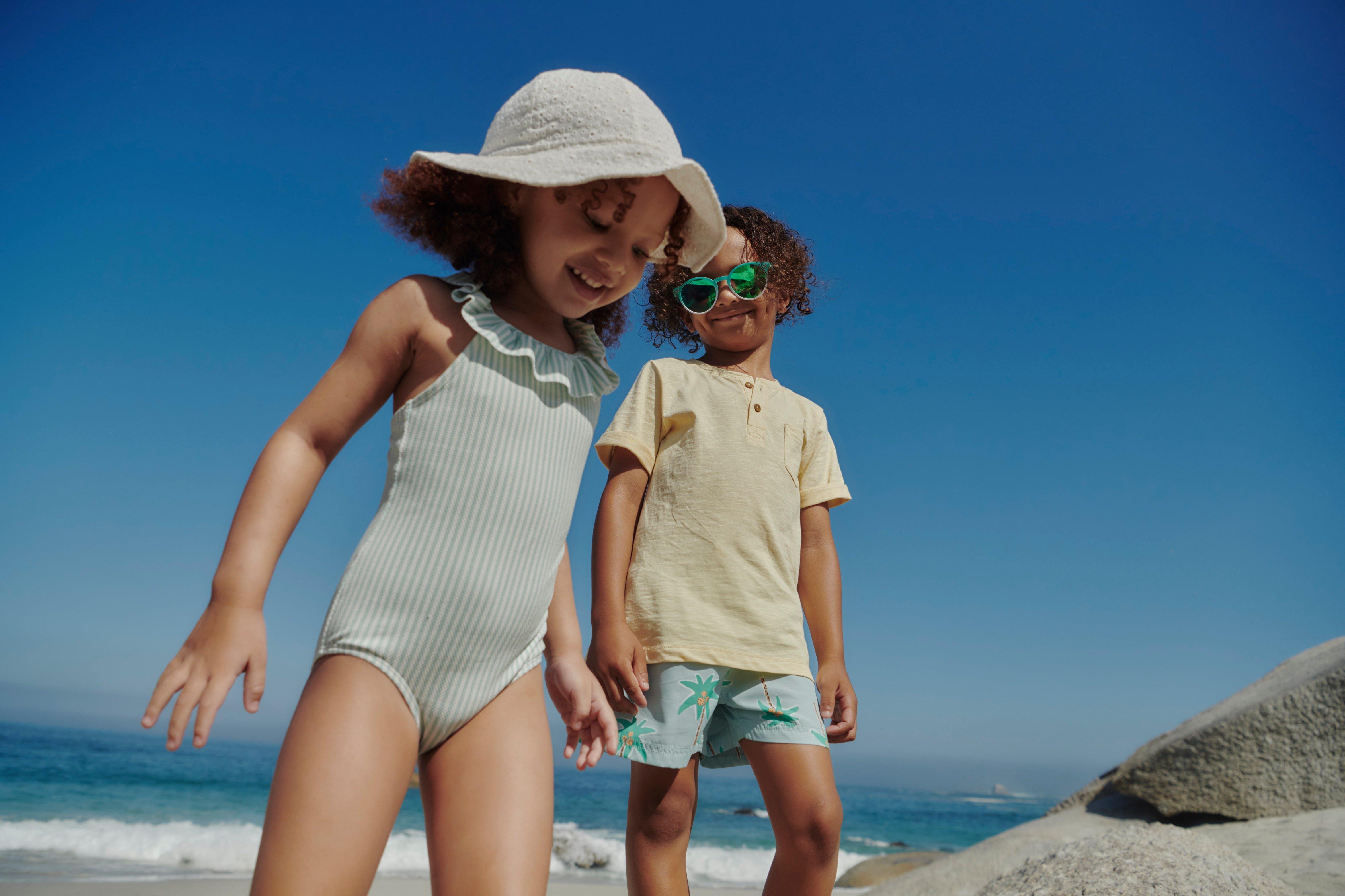 Dvě malé děti na pláži. Dívka v pruhovaném plavkách a slunečním klobouku a chlapec v lehkých šortkách a tričku kráčí vedle sebe.