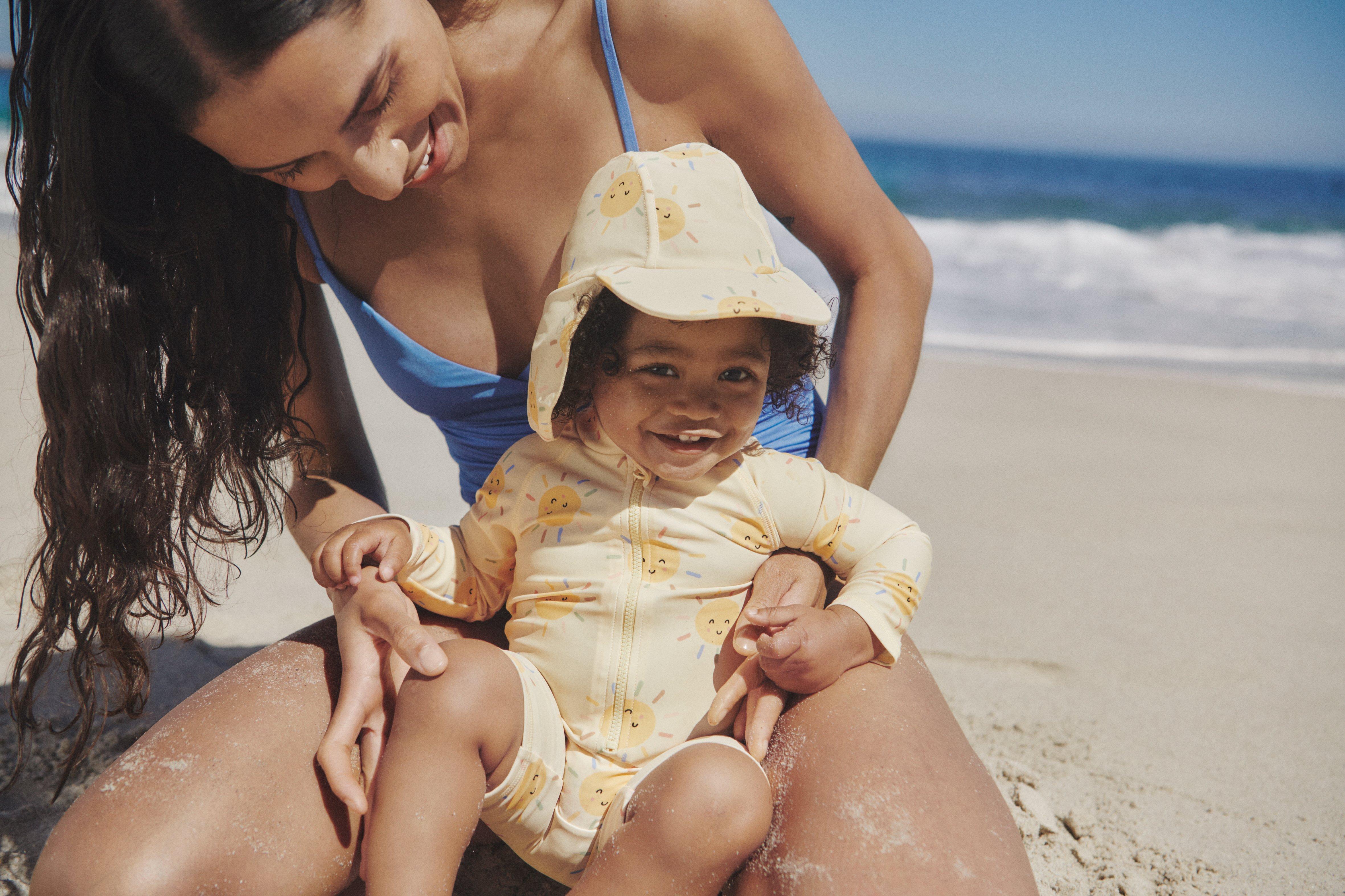  A mother sit down on the sand in a blue swimsuit. In her arms she holds a baby dressed in sun protection clothing.