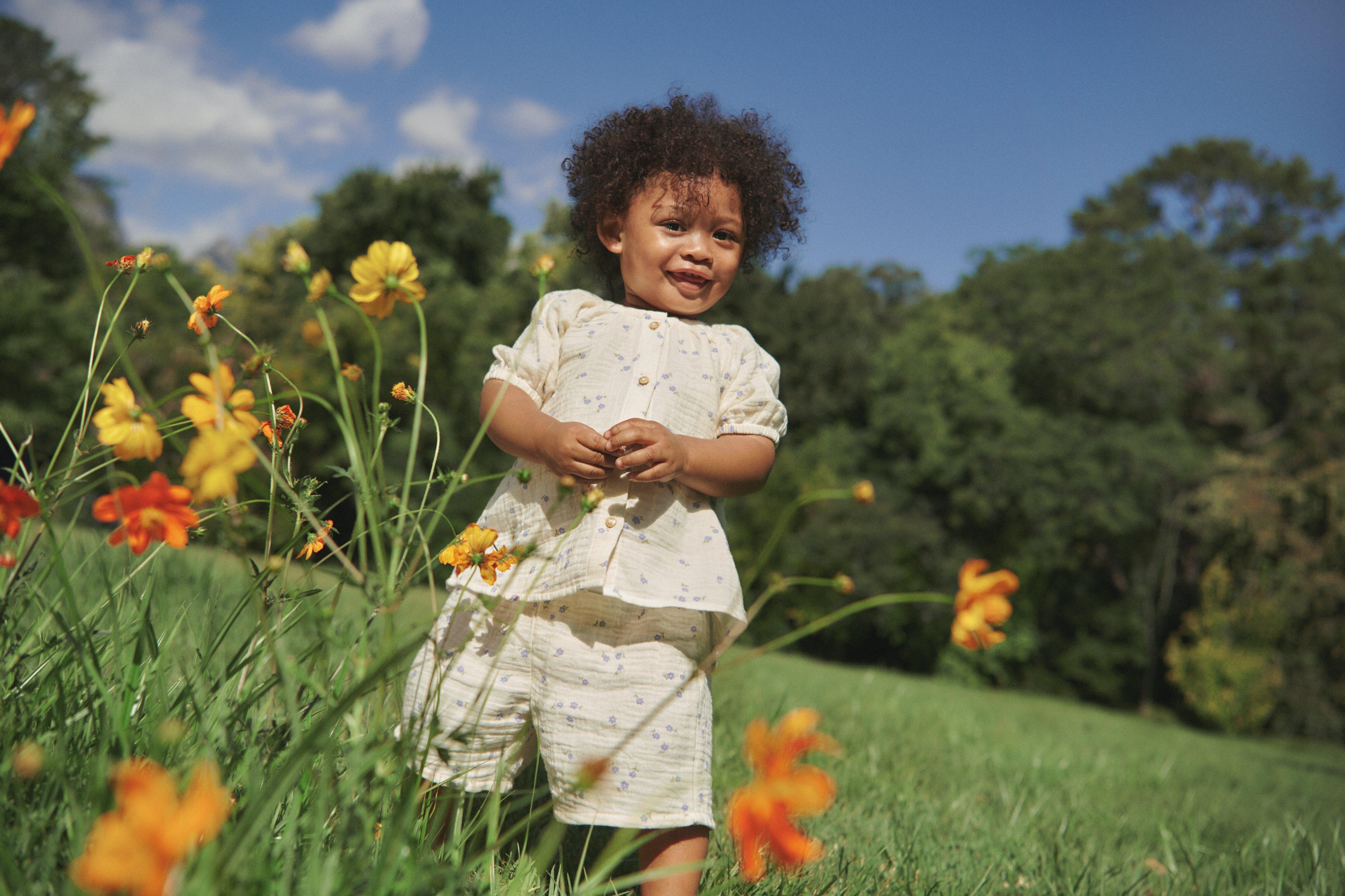 A little girl stands smiling in a flower meadow wearing a white blouse and relaxed shorts.