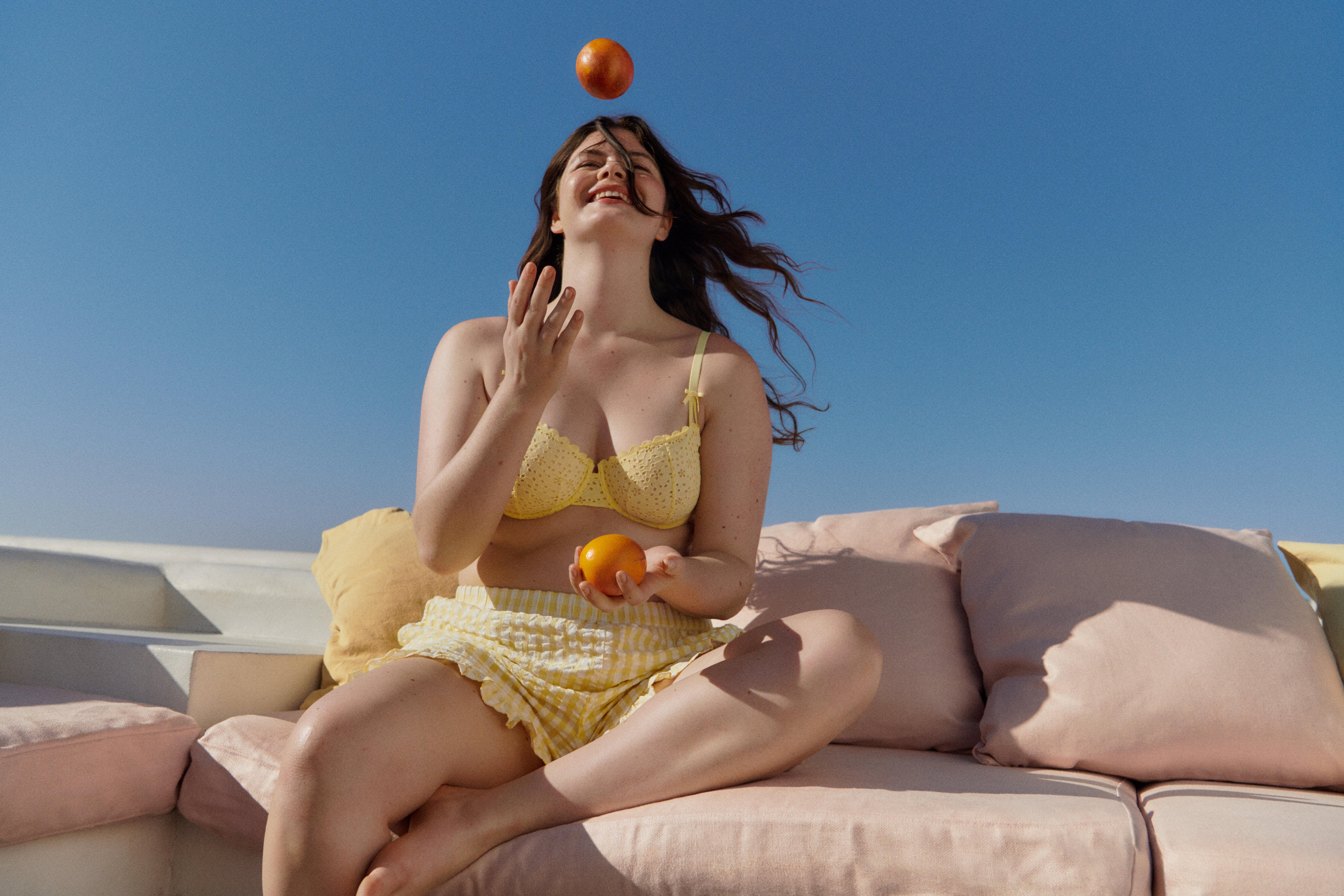 En leende kvinna sitter på en soffa utomhus och njuter av solen med ett äpple. Hon har på sig en gul bh och ett par gula shorts.
