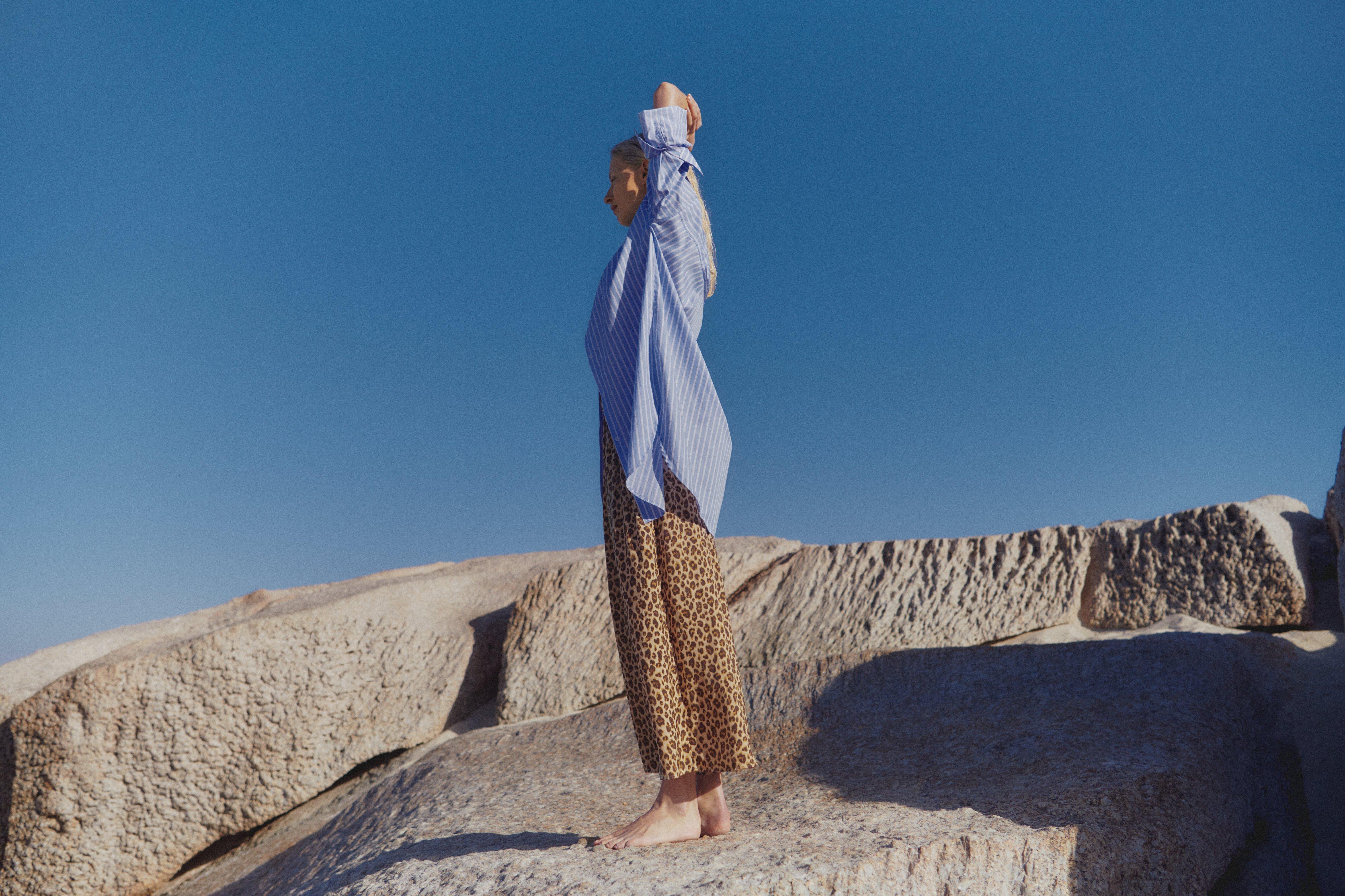 En kvinna som bär en lång blårandig skjorta och ett par vida byxor med leopardmönster ses från sidan. Hon står med armarna korsade över huvudet och tittar ut ur ramen.