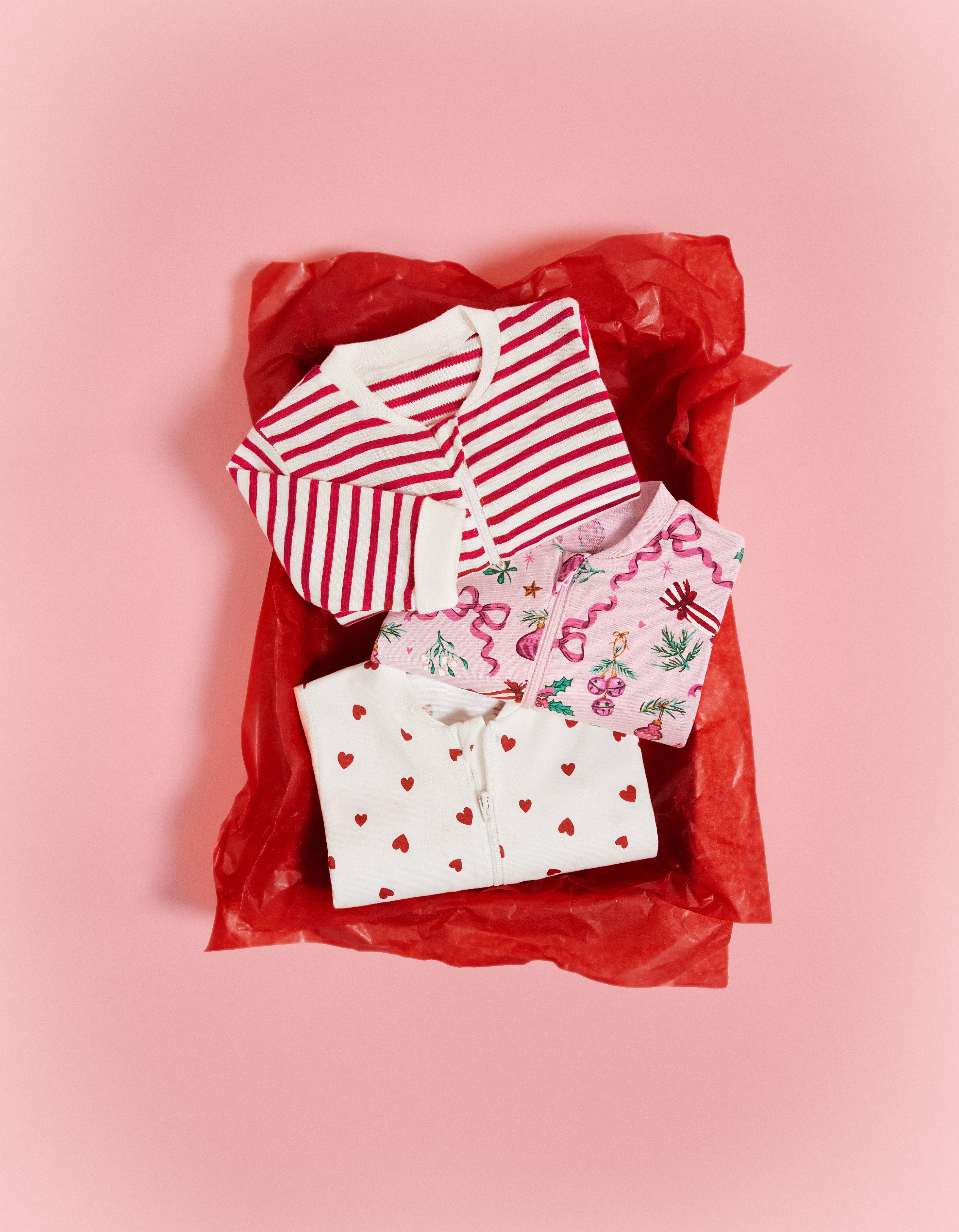 Tři pyžama pro miminka s vánočními vzory leží na červeném balicím papíru na růžovém stole.