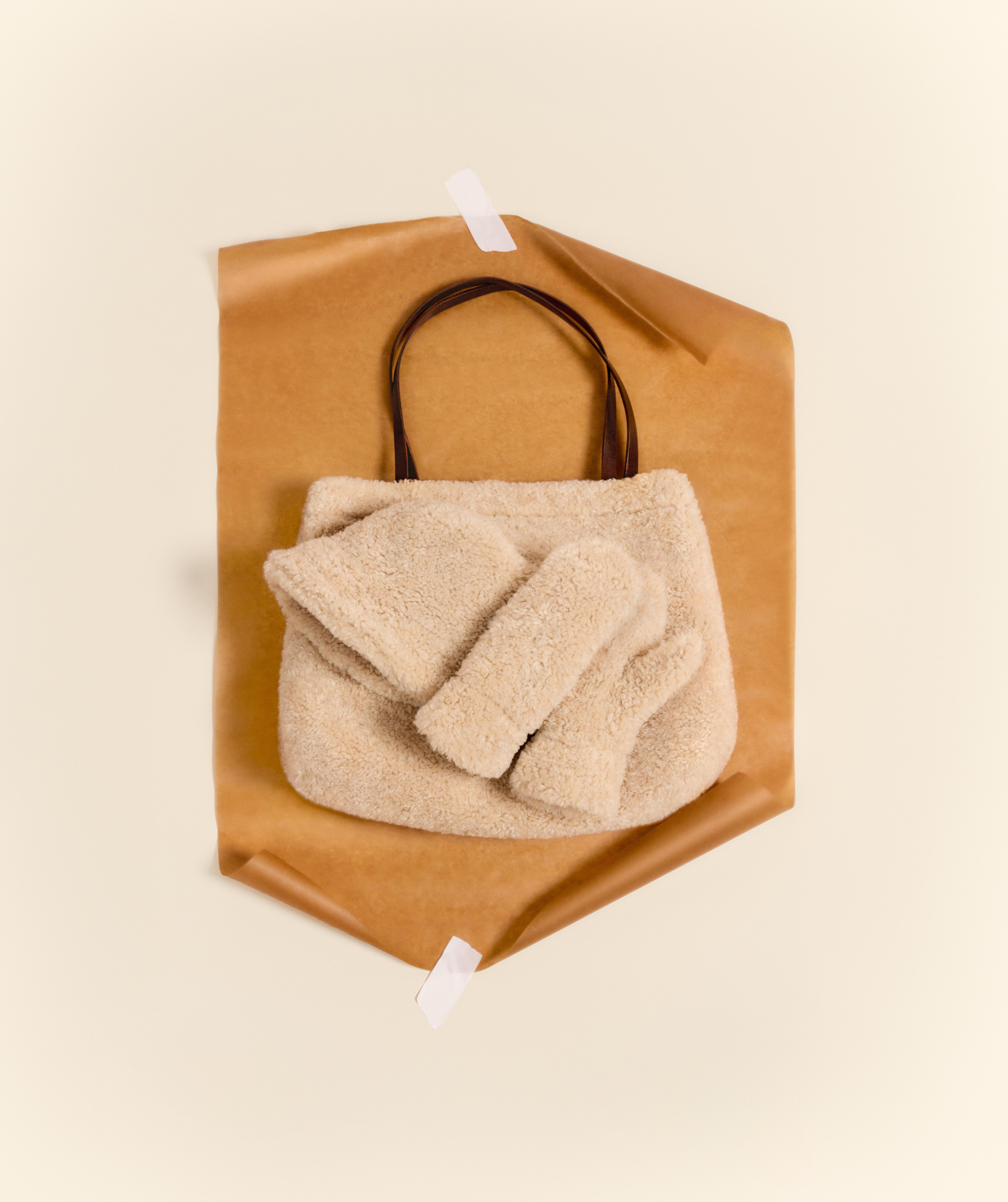 Čepice, pár palčáků a taška, vše v světle béžovém umělém kožíšku, leží na přírodně zbarveném balicím papíru připravené k zabalení.