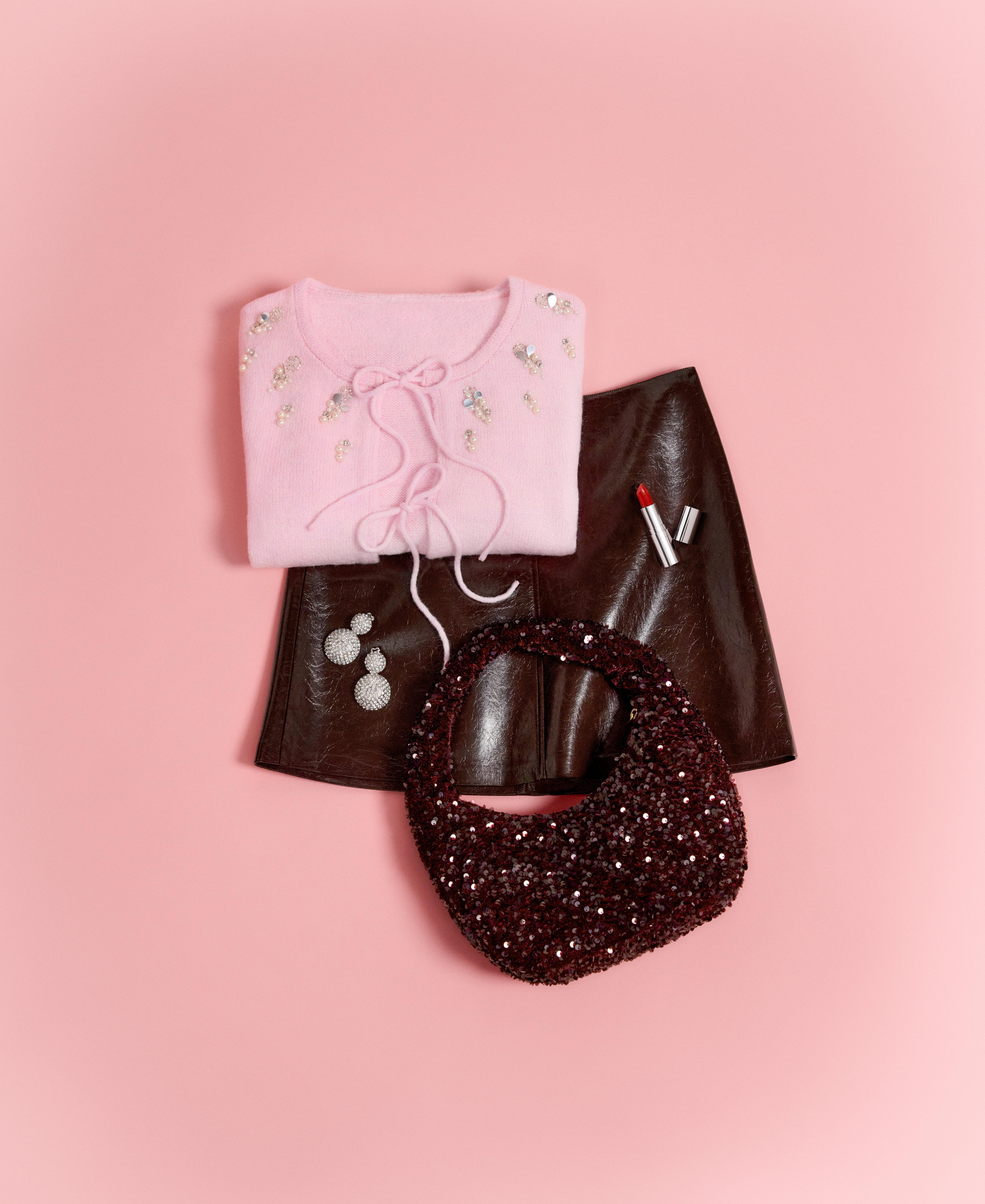 A close-up of a pink sweater, a faux leather skirt, makeup and a bag lying on a pink background.