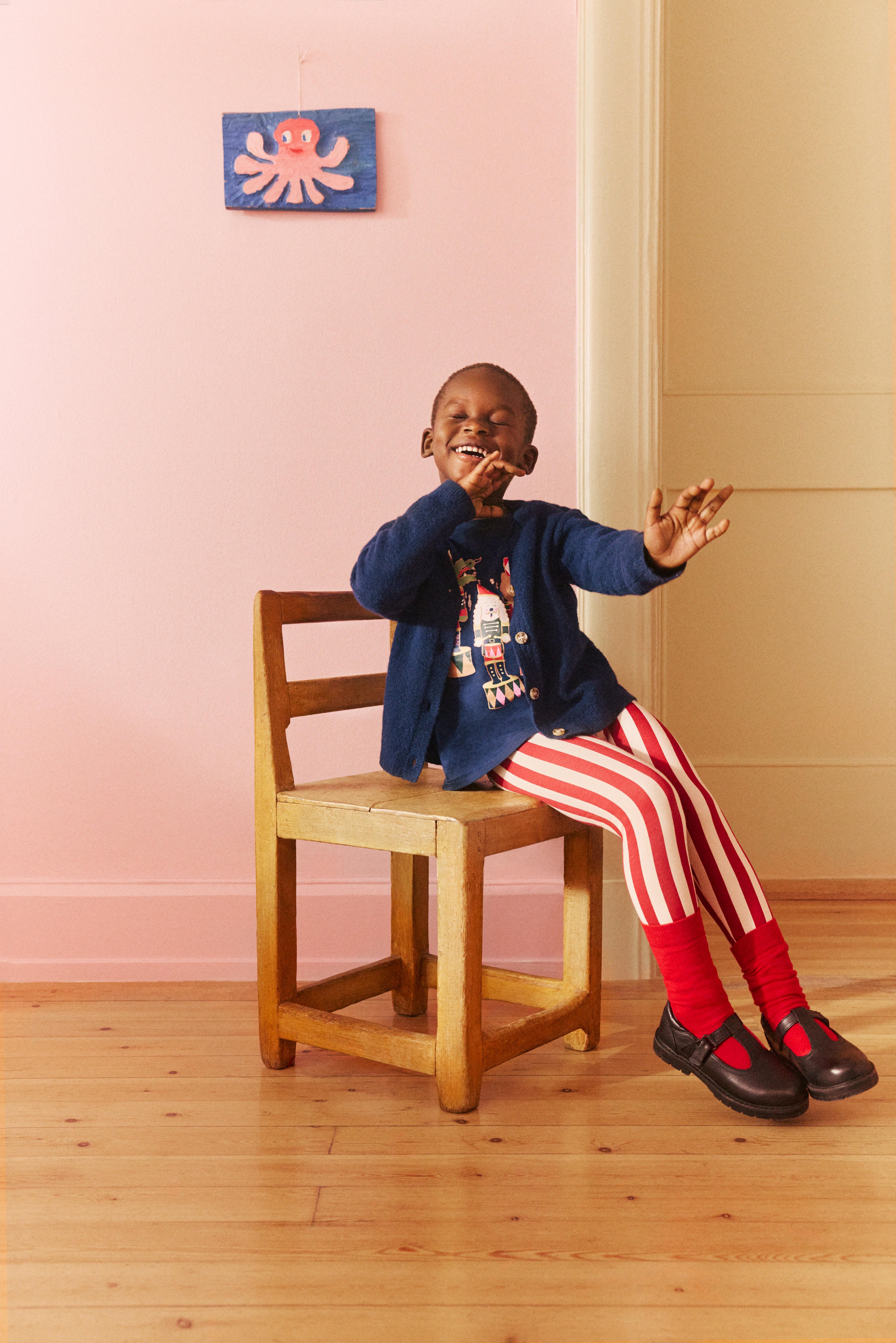 Dítě sedí na dřevěné židli a zdá se, že hraje na dechový nástroj. Dítě má na sobě tričko s potiskem kresleného zvířecího orchestru.