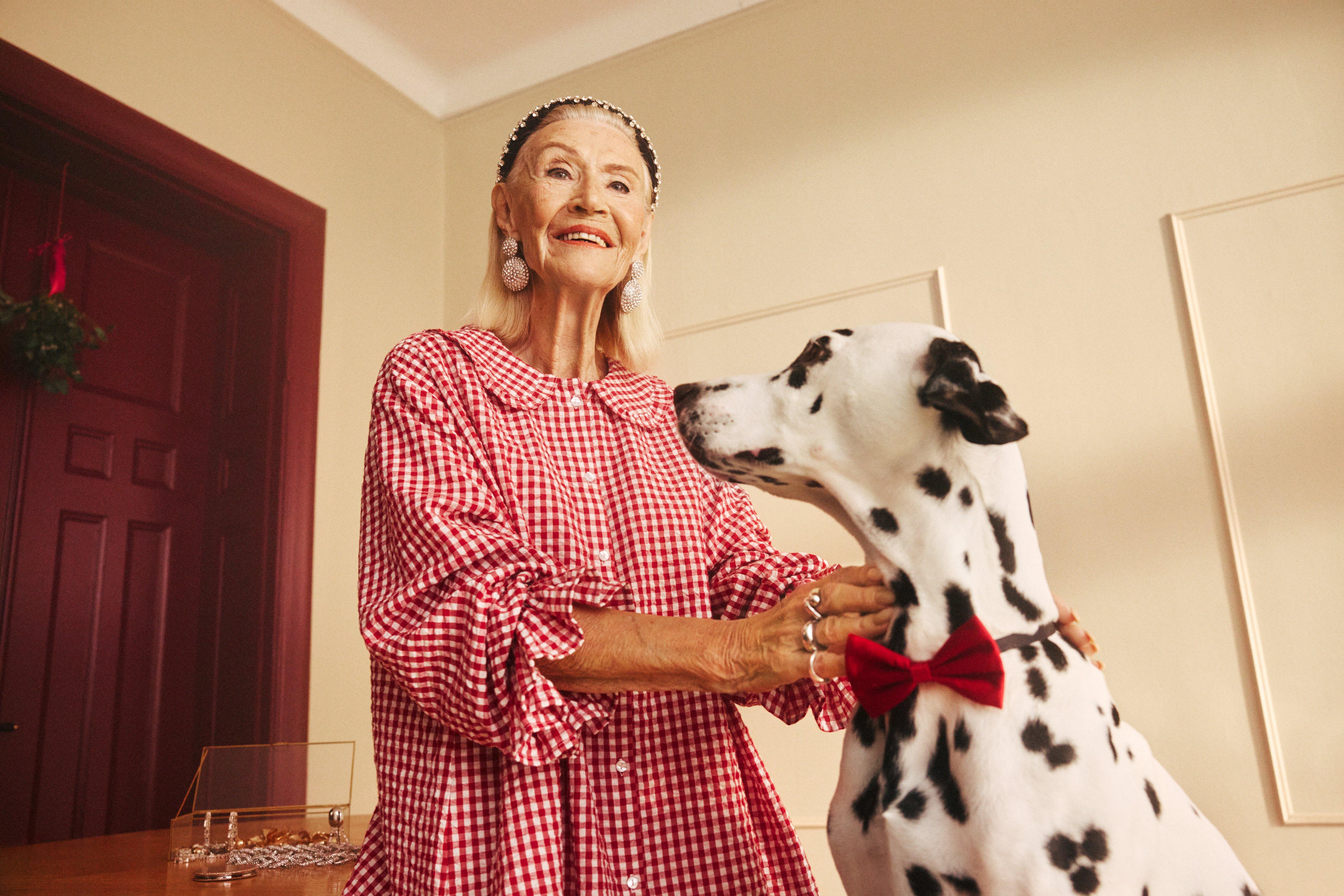 Žena s líčením, oblečená v červené kostkované pyžamové košili s brookovým vzorem, s límečkem a dlouhými rukávy s volánky, stojí v místnosti s dalmatinem po svém boku.