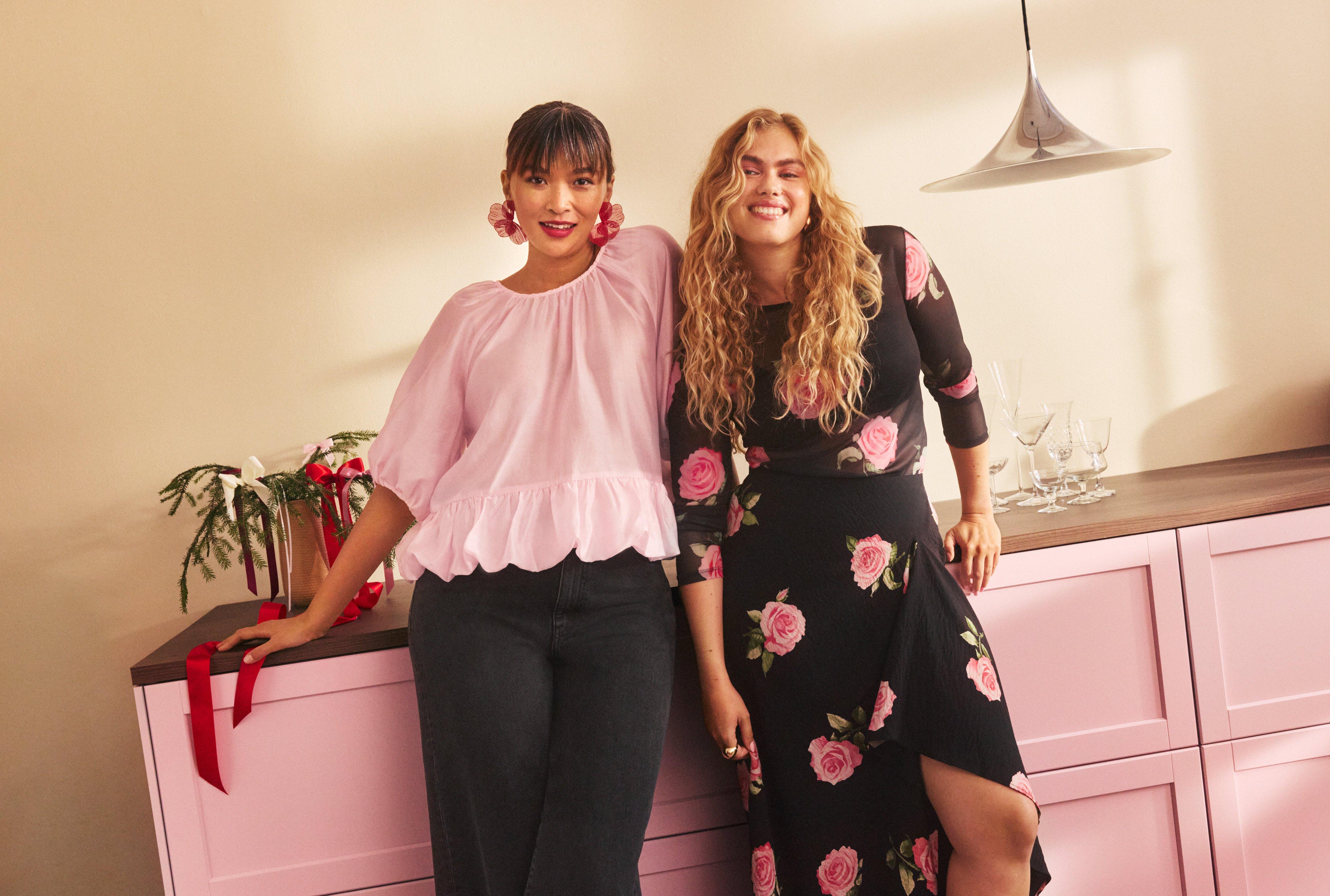 Two smiling women. One in a black skirt and top with roses. One in a pink blouse and black pants.