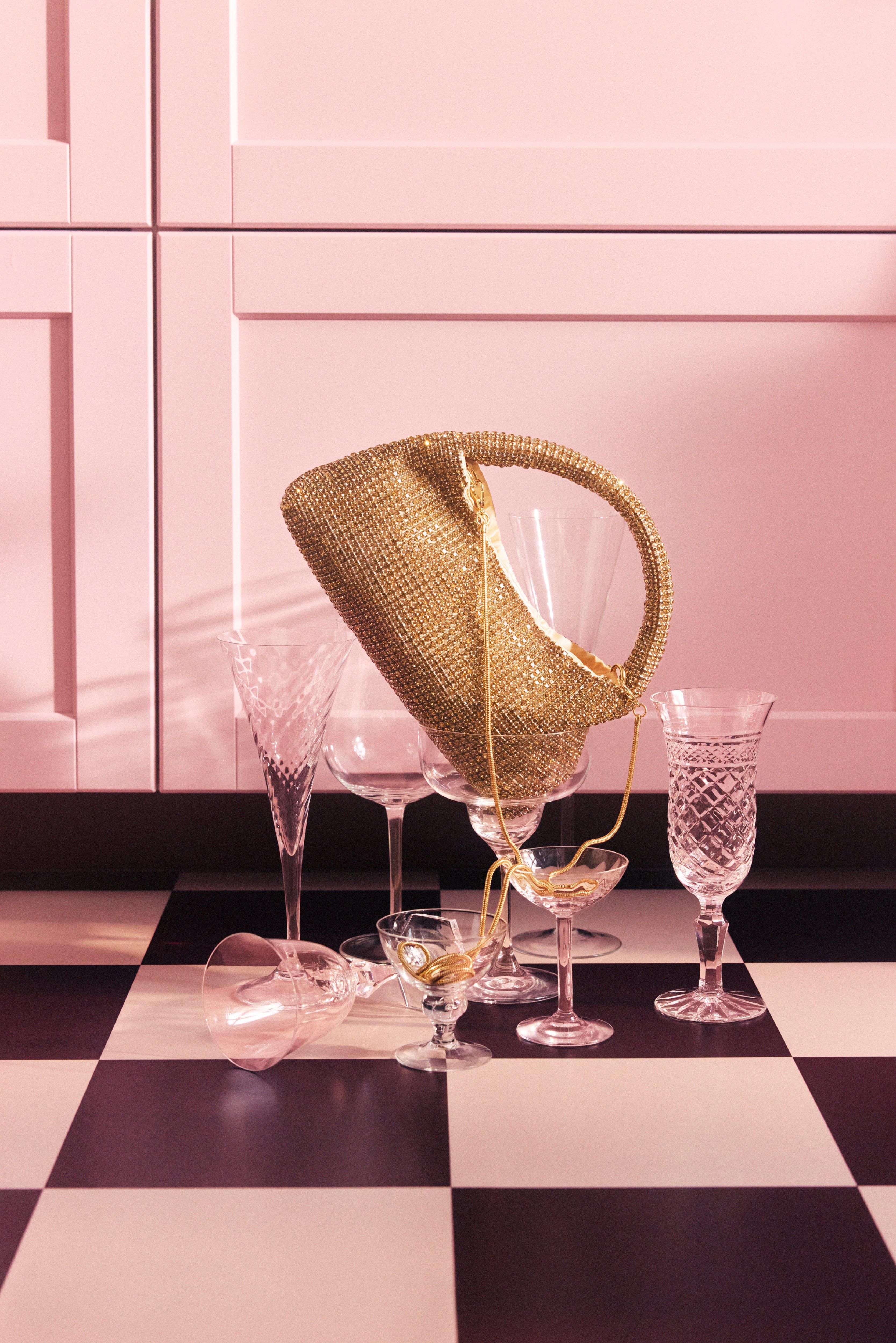 A gold bag with rhinestones is leaning in a glass that stands on a checkered floor. Next to it are several glasses in different shapes and sizes.