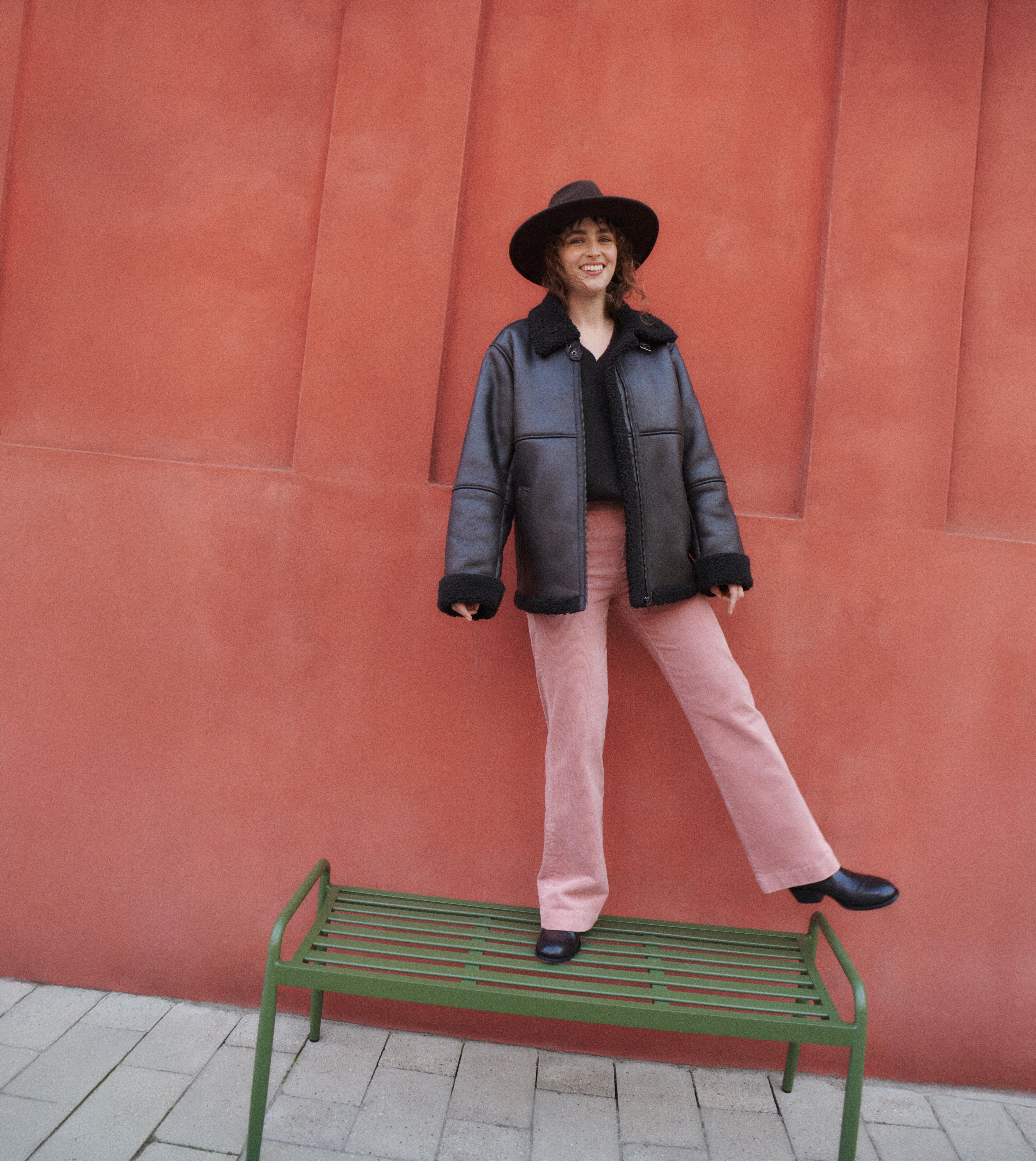  A woman in a pair of beige pants, a faux leather jacket with fur, and a wide-brimmed hat stands on one leg on a bench and balances. She smiles broadly at the camera.