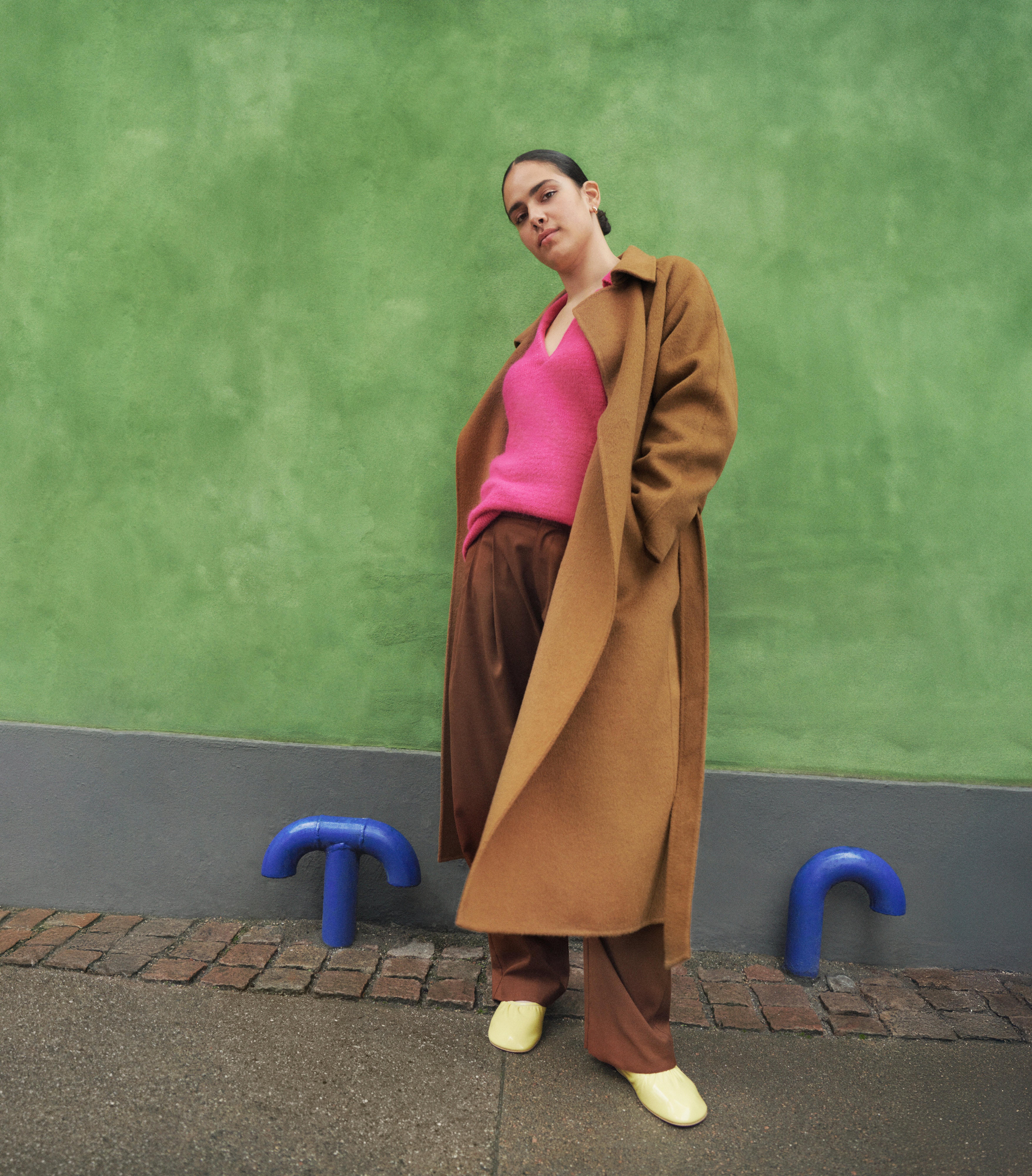  A woman wearing a long brown coat, pink sweater and a pair of brown pants stands against a green wall.