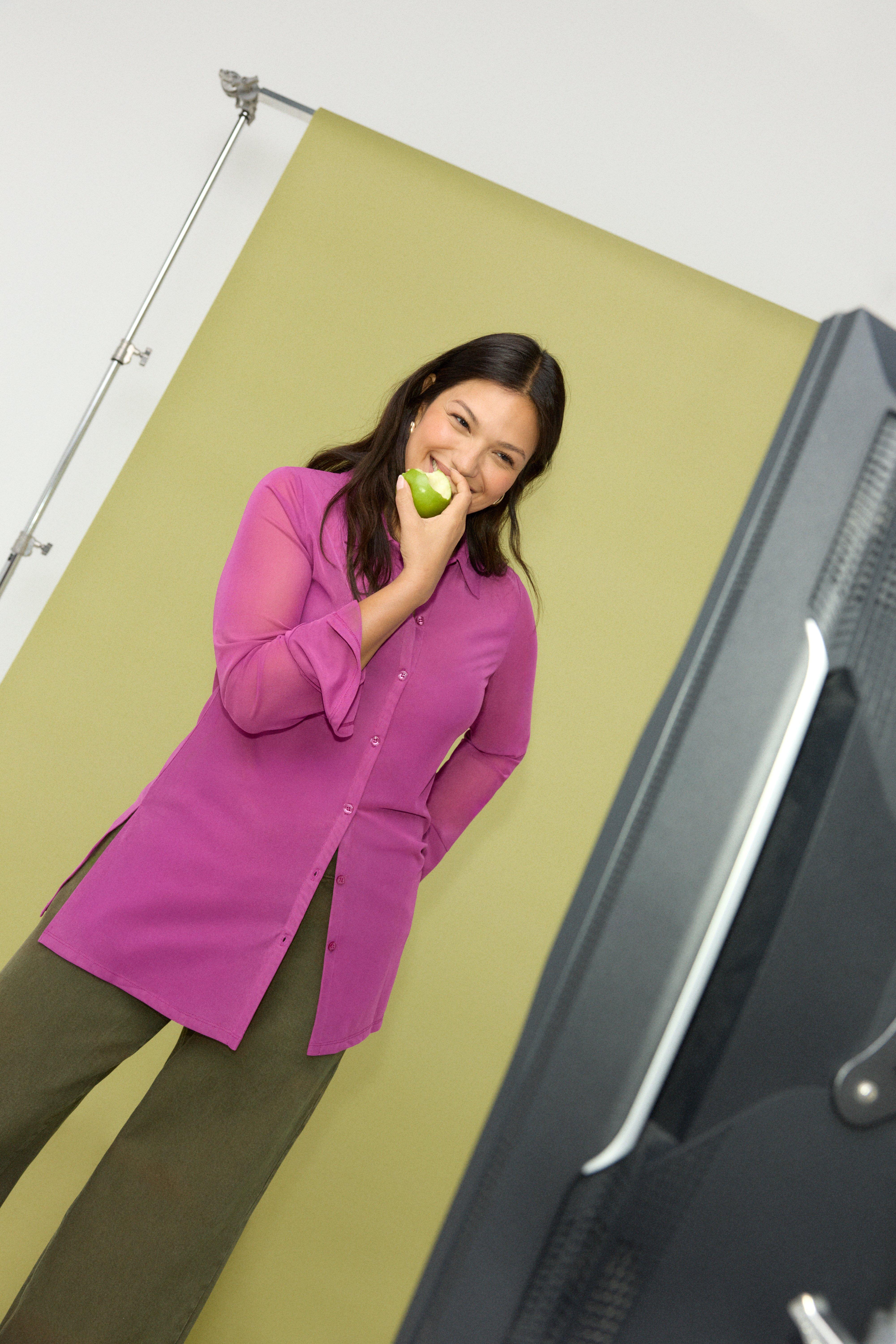 A woman wearing a purple long jersey shirt with collar and long sleeves and a pair of green pants looks into the camera and laughs. She holds a green apple in her hand.
