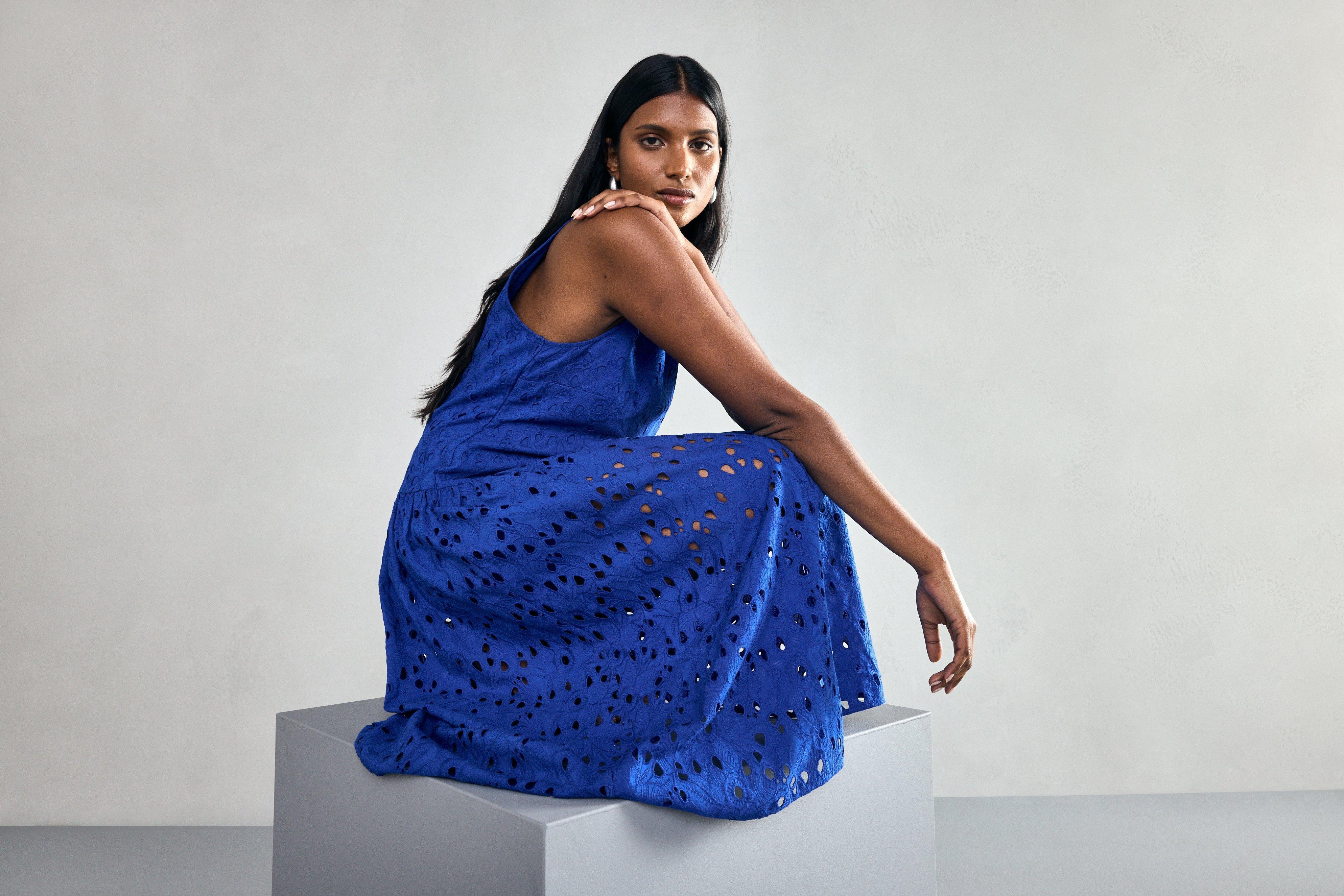  A woman is squatting wearing a blue sleeveless midi dress with a low, gathered waist and broderie anglaise details.