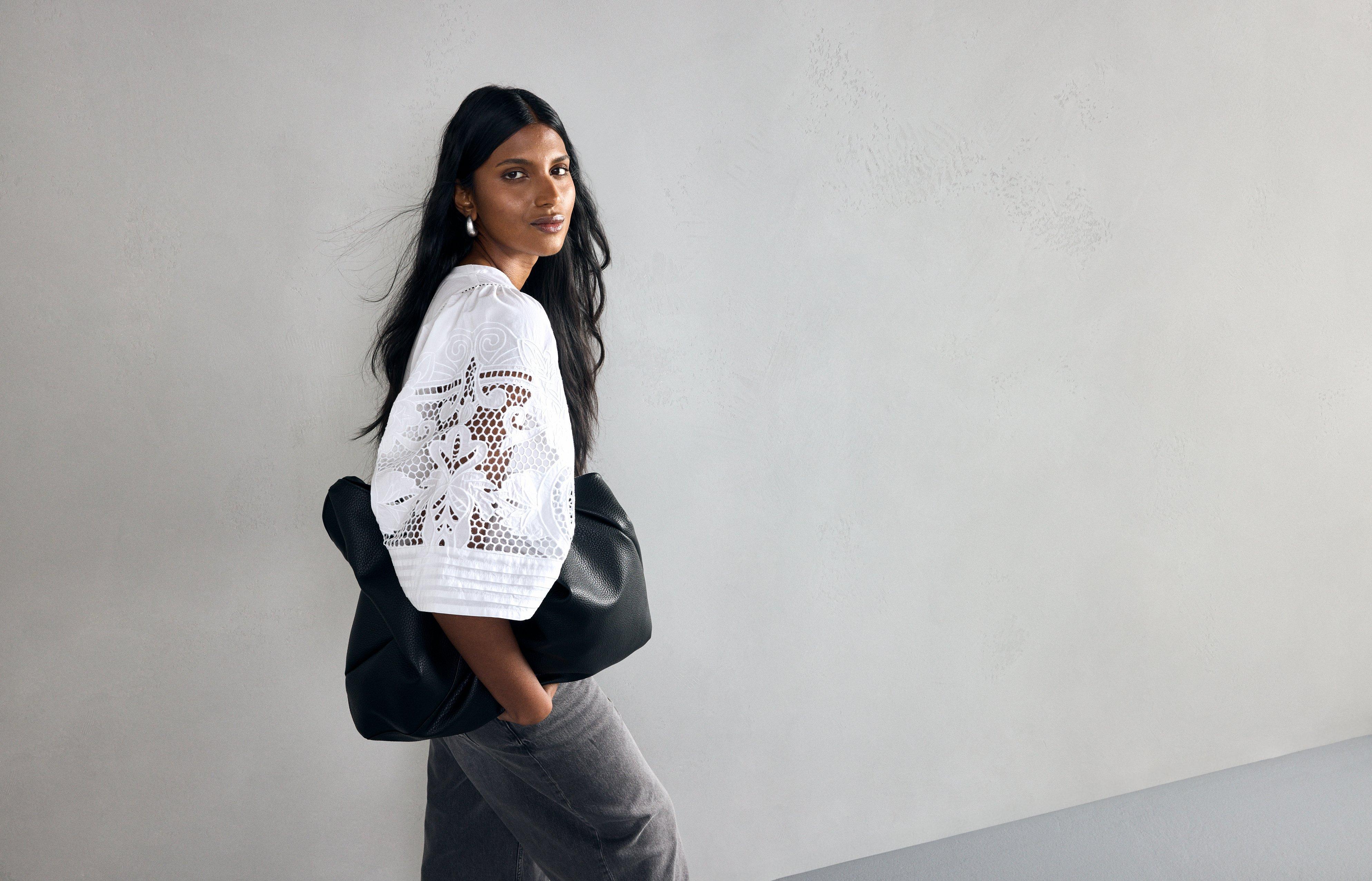  A woman wearing denim shorts and a white woven blouse in crisp cotton with a relaxed fit and wide cropped sleeves with embroidered details. The woman is holding a large bag.