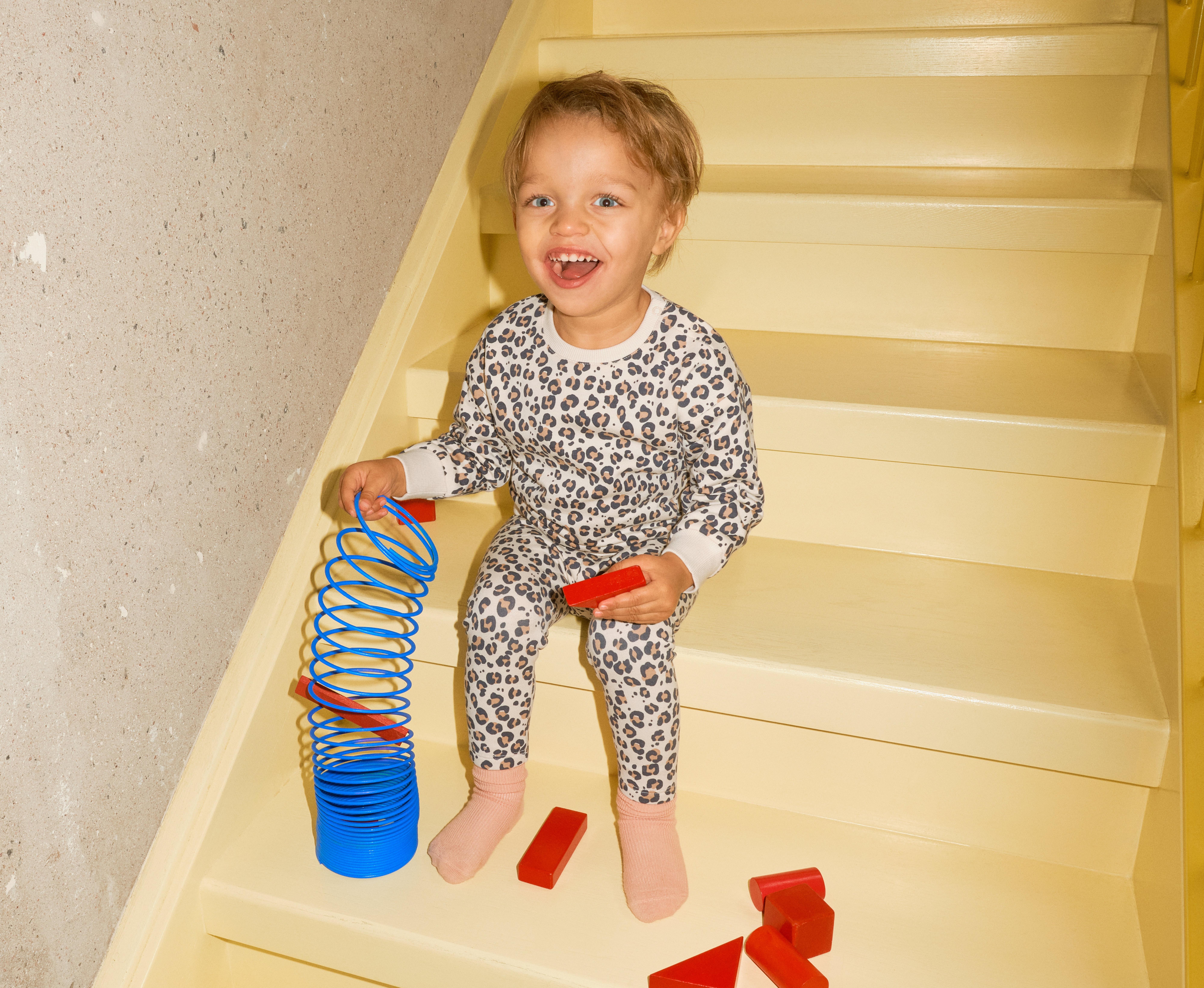 Ett barn klädd i en ett med djurprint sitter i en trappa och leker. Barnet ler mot kameran.