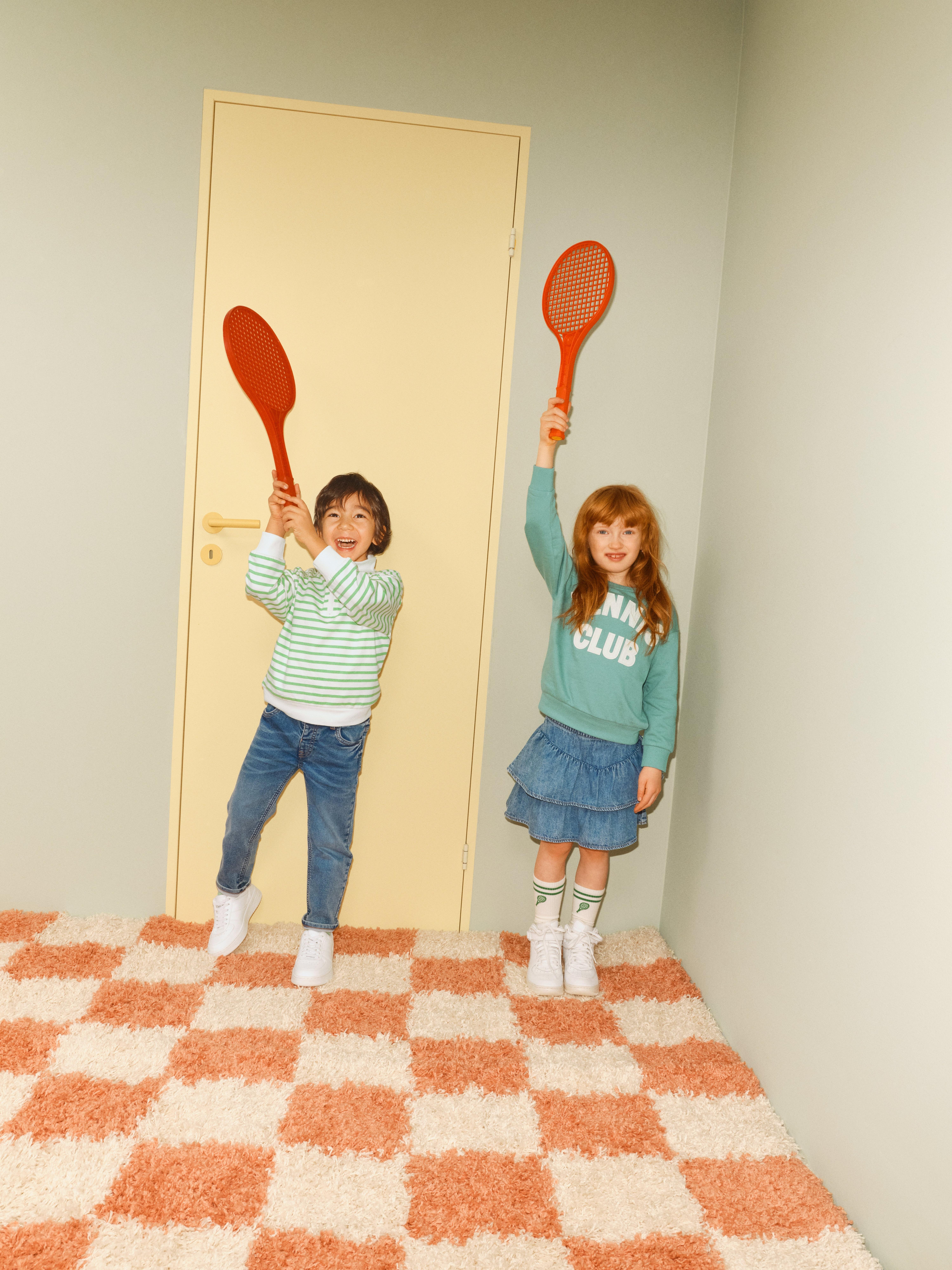 Två barn står på ett rutigt golv och håller upp varsin tennis racket i luften.