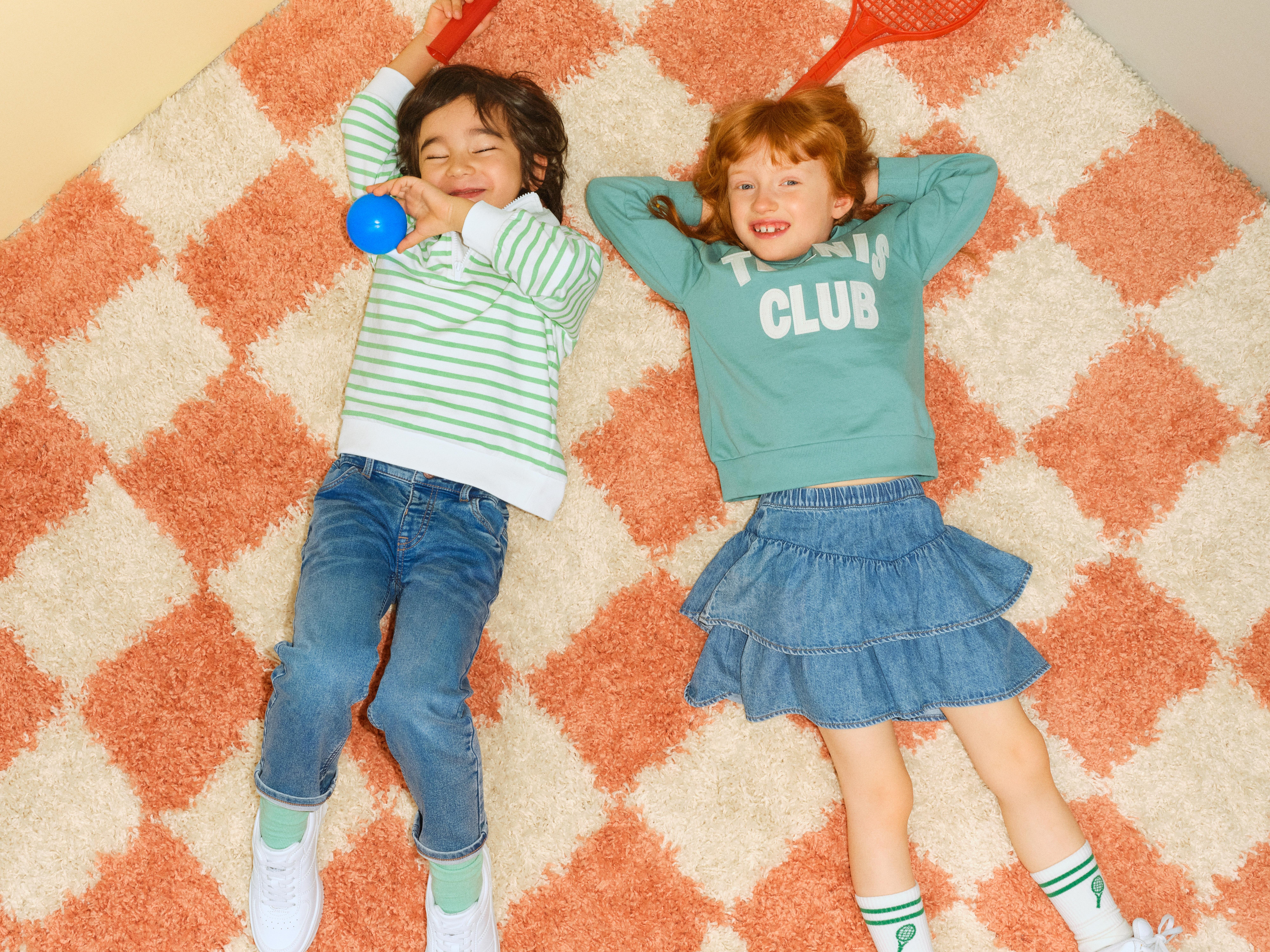 En liten tjej i grön tröja och jeanskjol och en liten kille i jeans och randig tröja ligger på ett rutigt golv och ser ut mot kameran.