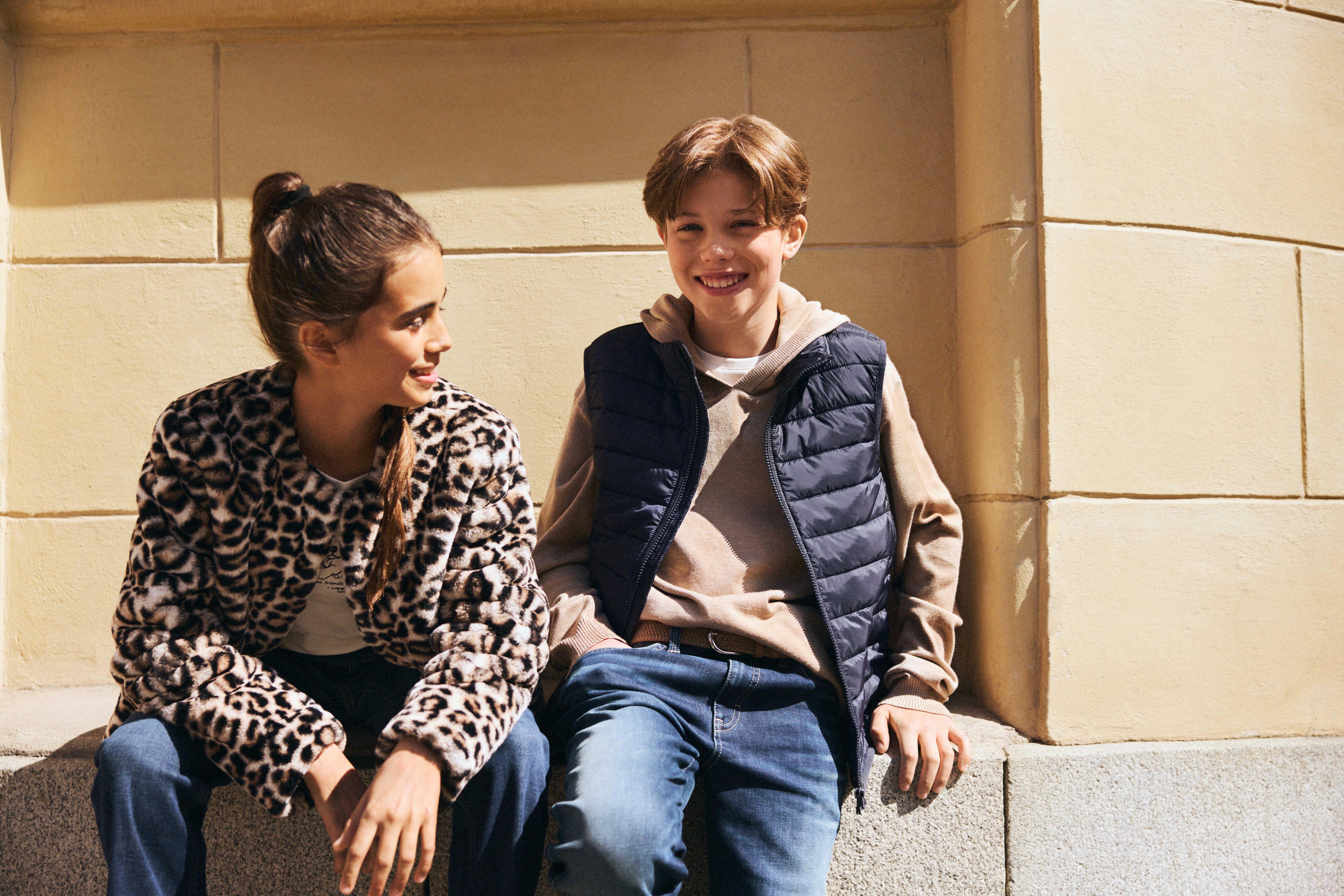 Dívka a chlapec sedí před žlutou kamennou fasádou. Dívka se dívá na chlapce, který se usmívá do kamery. Dívka má na sobě kabát z umělé kožešiny a džíny. Chlapec má na sobě džíny, šedou mikinu a prošívanou vestu.