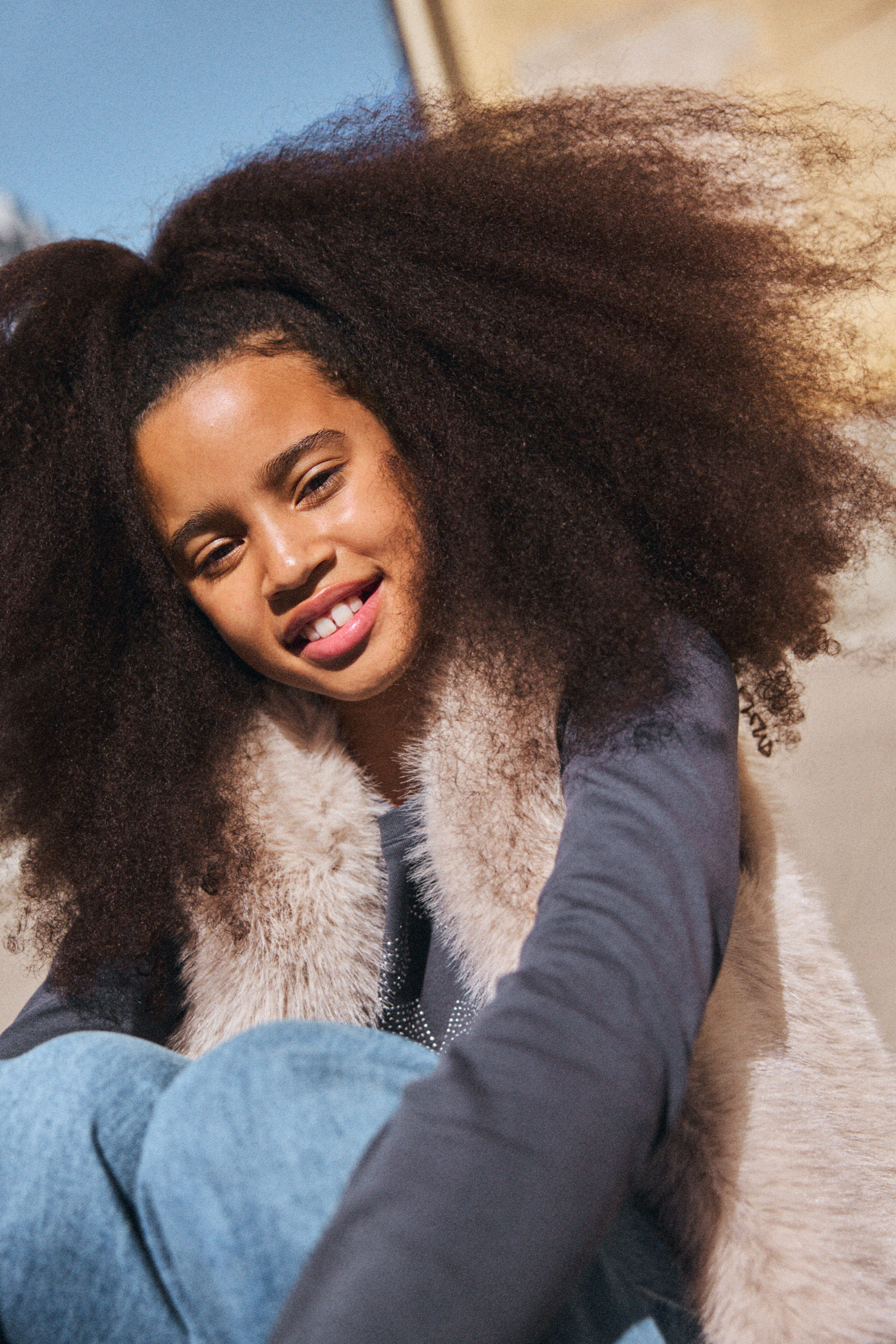 Detailní záběr usmívající se dívky sedící a dívající se do kamery. Má na sobě džíny, šedý svetr a vestu z umělé kožešiny.