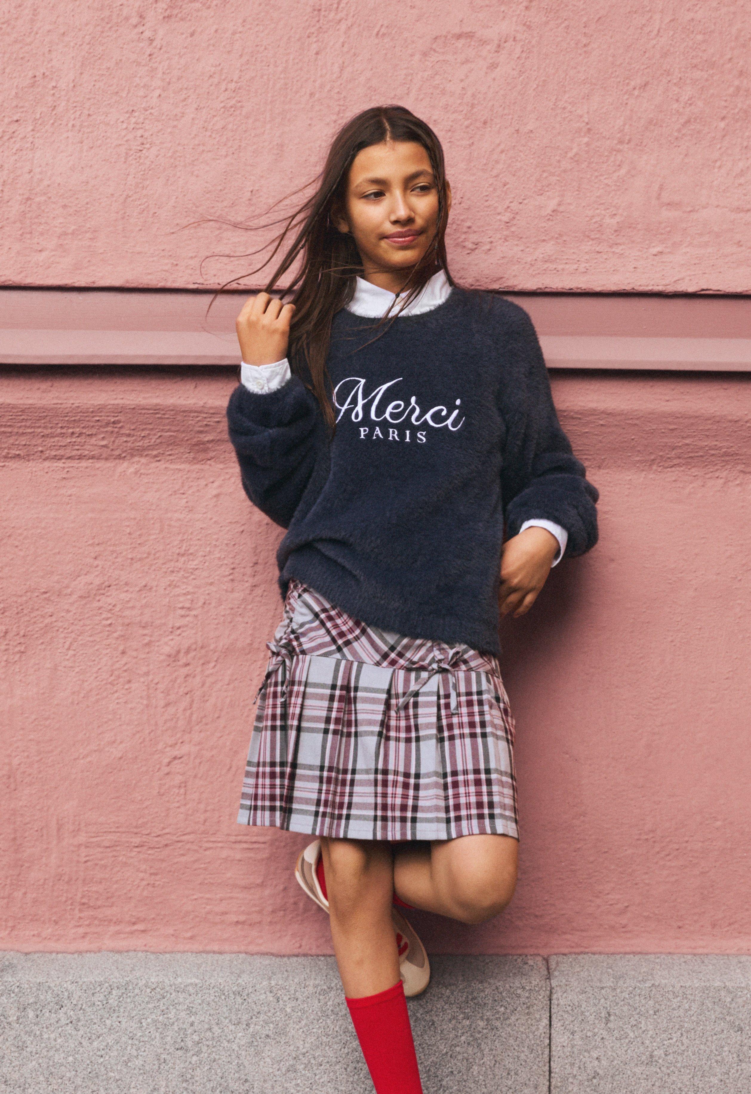 Školní dívka stojí opřená o růžovou kamennou zeď. Má jednu ruku ve vlasech. Na sobě má kostkovanou sukni a tmavý college svetr s potiskem.
