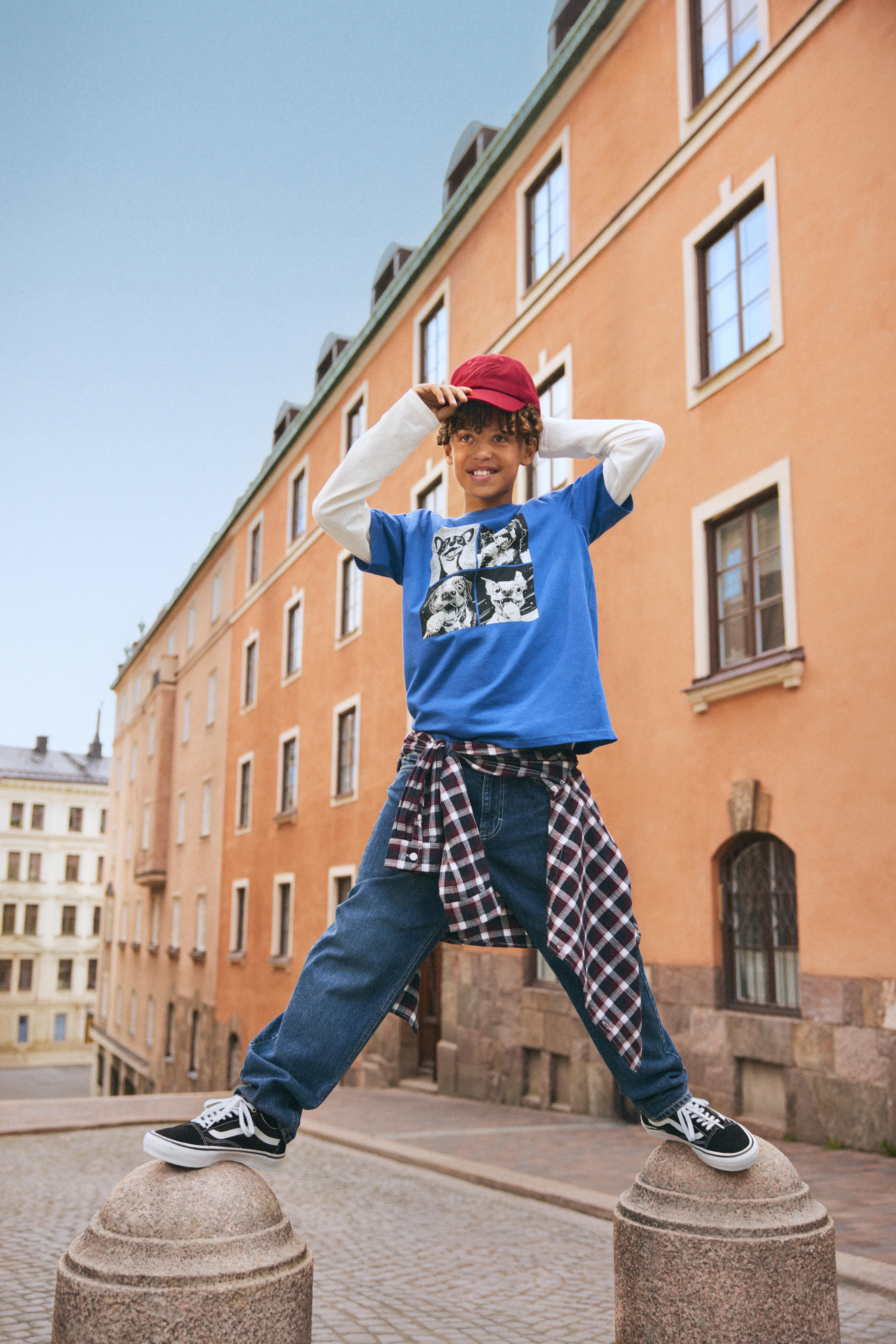 Chlapec stojící na dvou kamenných sloupech v městské oblasti, nosící červenou čepici, modré tričko s grafikou tří koček, bílá košile s dlouhými rukávy pod ním, volné modré džíny, černobílé tenisky a kostkovanou košili uvázanou kolem pasu. Za ním je oranžová budova s několika okny.
