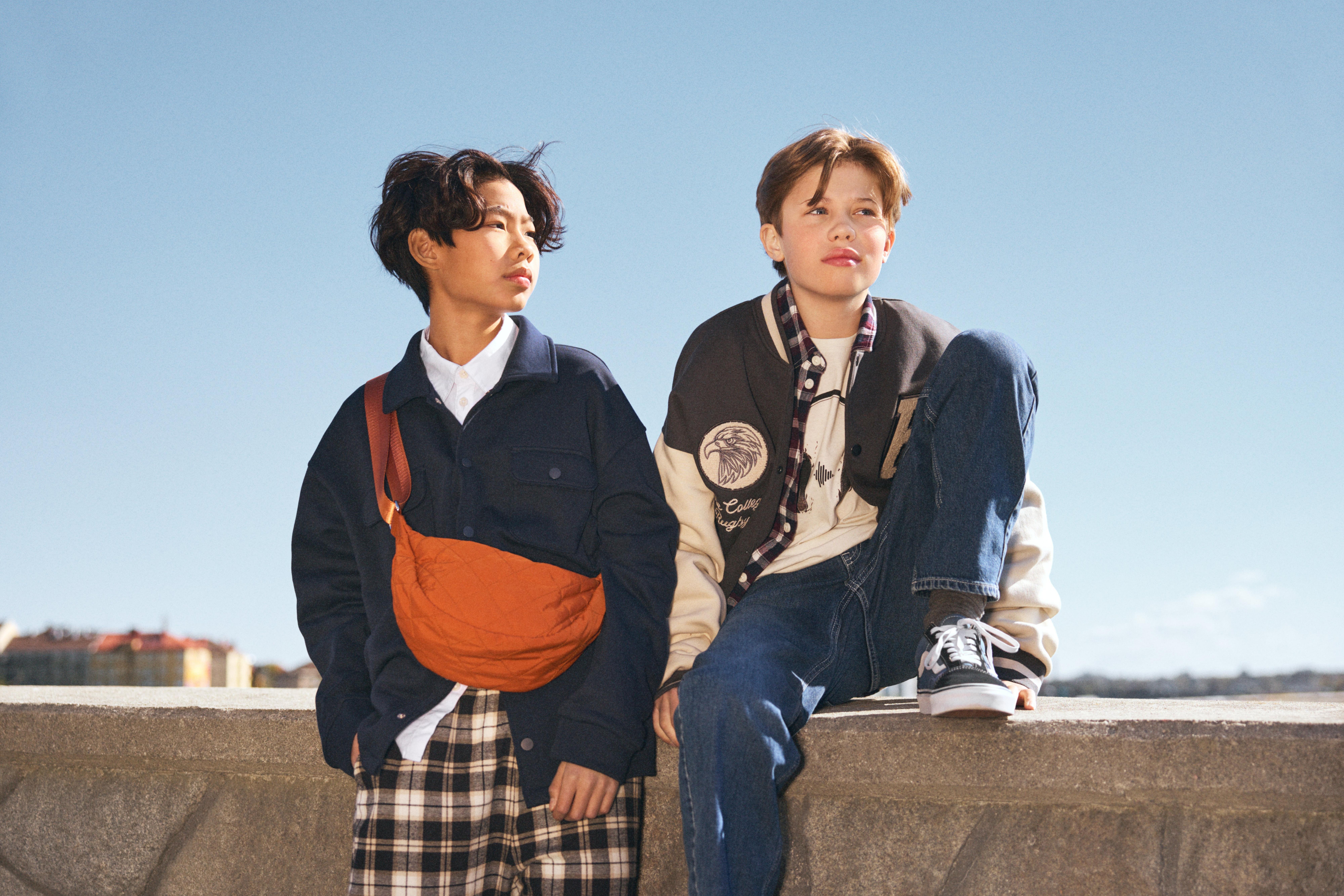 Dva kluci u kamenné zdi s modrou oblohou v pozadí. Jeden kluk se opírá o zeď, má na sobě tmavě modrou košili, bílou košili a kostkované kalhoty. Přes rameno má oranžovou tašku. Druhý kluk sedí na zdi. Má na sobě džíny, bílé tričko, kostkovanou košili a baseballovou bundu.