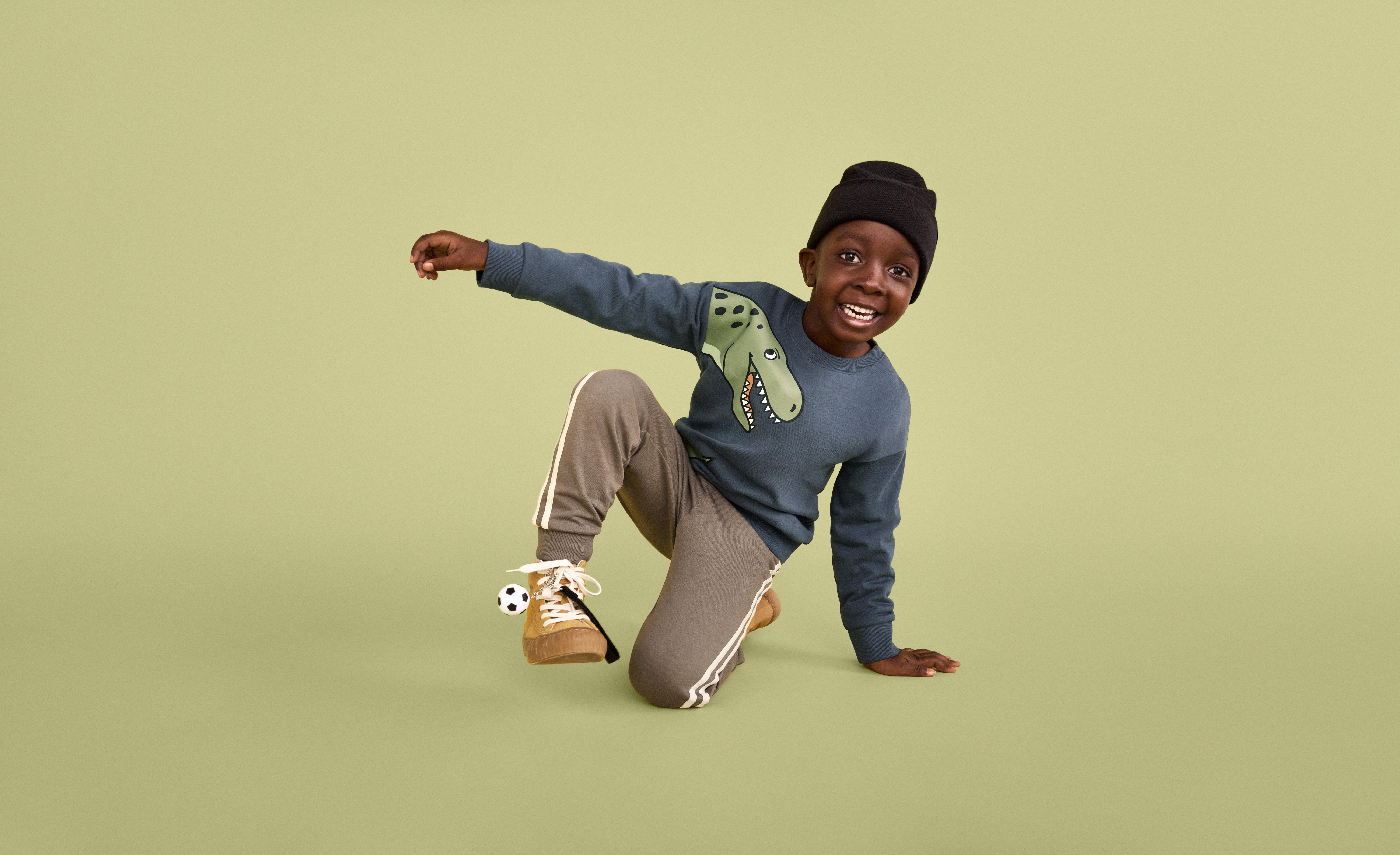 Chlapec oblečený v světle zelených teplácích s bílými pruhy po nohavicích, zeleném tričku s dinosaurem a tmavě zelené čepici se zdá, že breakdancuje. Pozadí a podlaha jsou světle zelené.