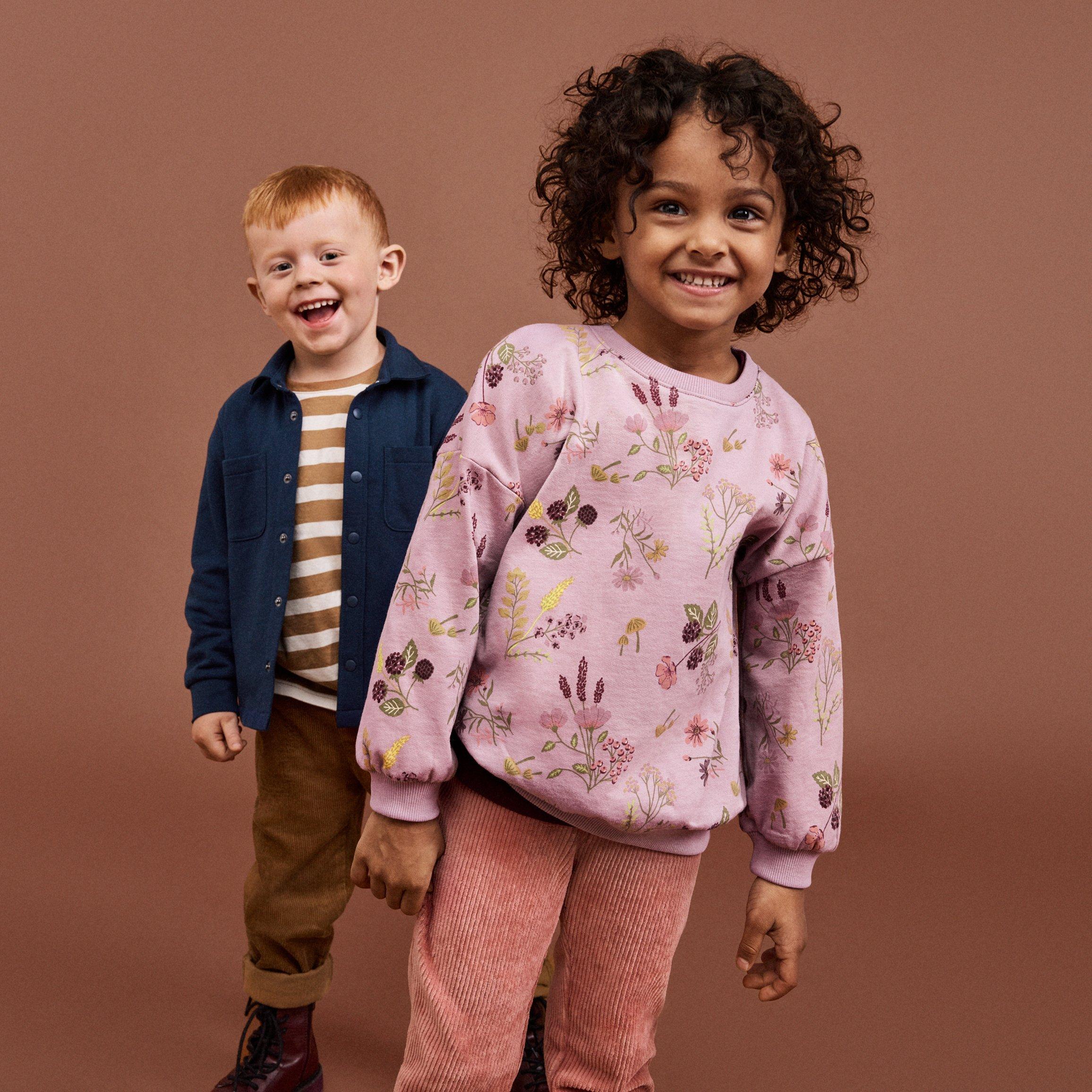 Dvě předškolní děti stojí proti hnědému pozadí. Obě se dívají do kamery a usmívají se. Vepředu je dívka v růžovém květinovém tričku a růžových kalhotách. Za ní je chlapec v pruhovaném tričku, modré košili a hnědých kalhotách.