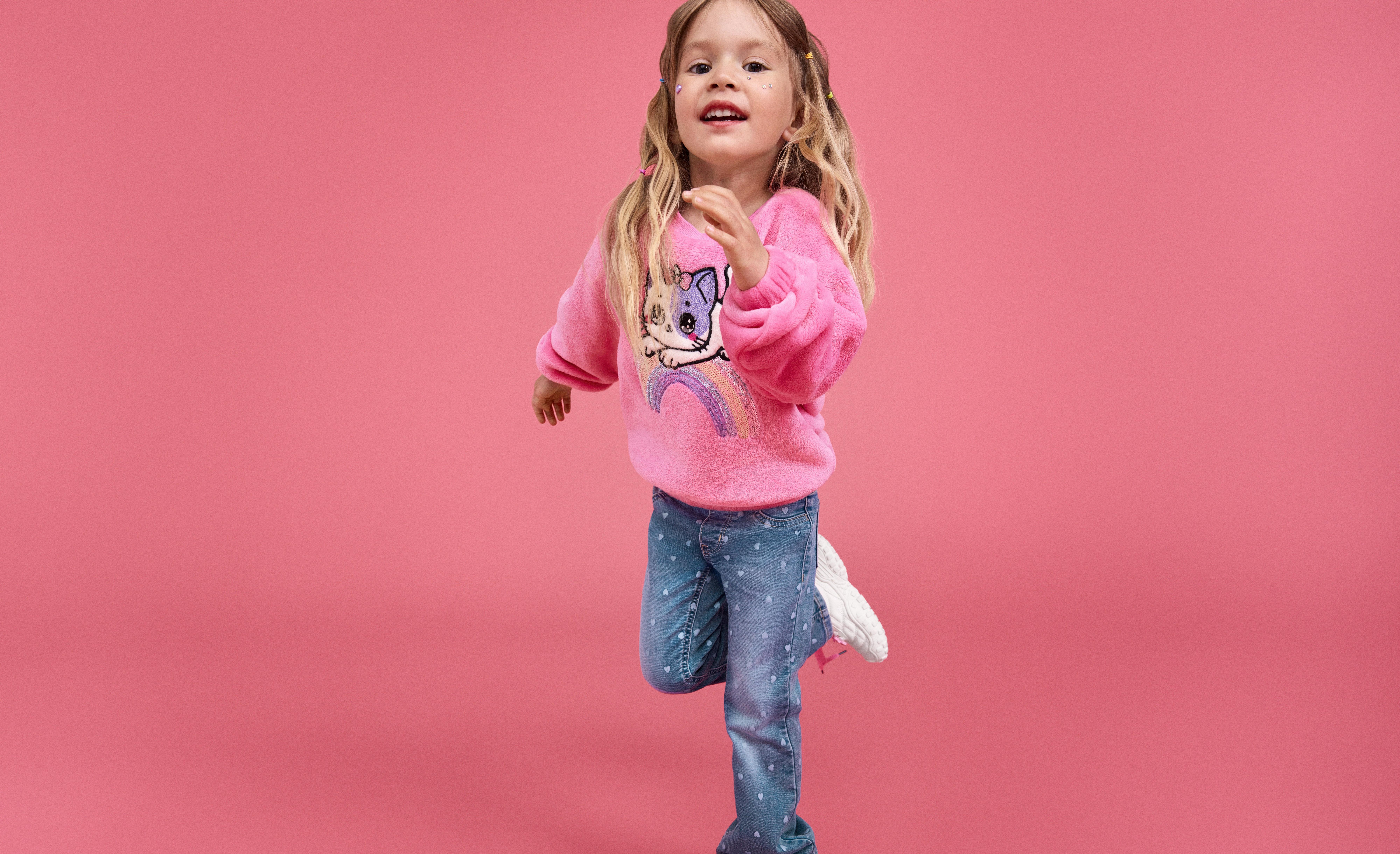 Dívka v růžovém svetru s kreslenou kočkou a modrých džínách s bílými vzory stojí proti jednobarevnému růžovému pozadí.