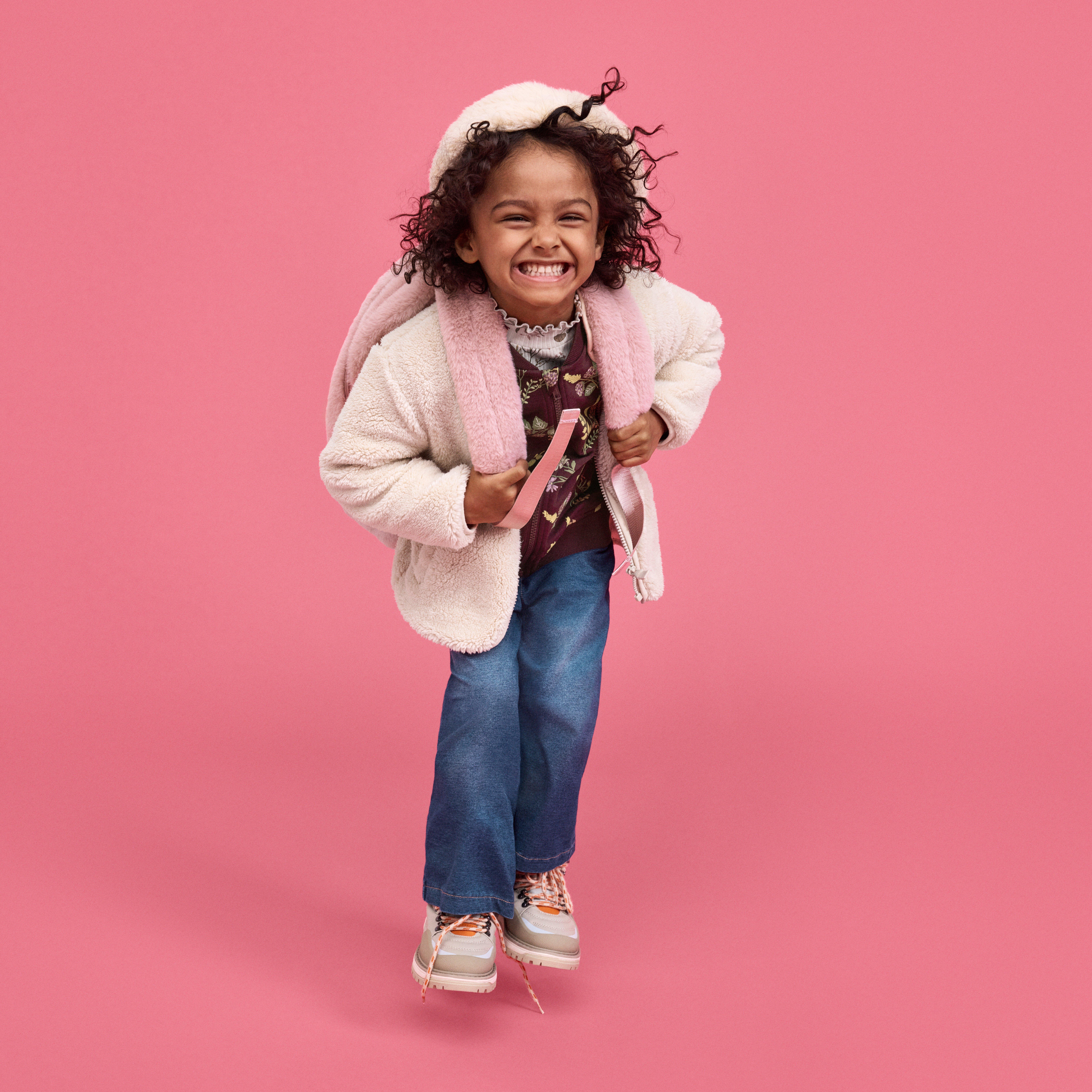 Dívka s kudrnatými vlasy, oblečená v světle růžové bundě s kapucí, modrých džínách a bílých teniskách s růžovými akcenty, stojí na jednobarevném růžovém pozadí. Dívka drží popruhy světle růžového batohu, který má na obou ramenech.