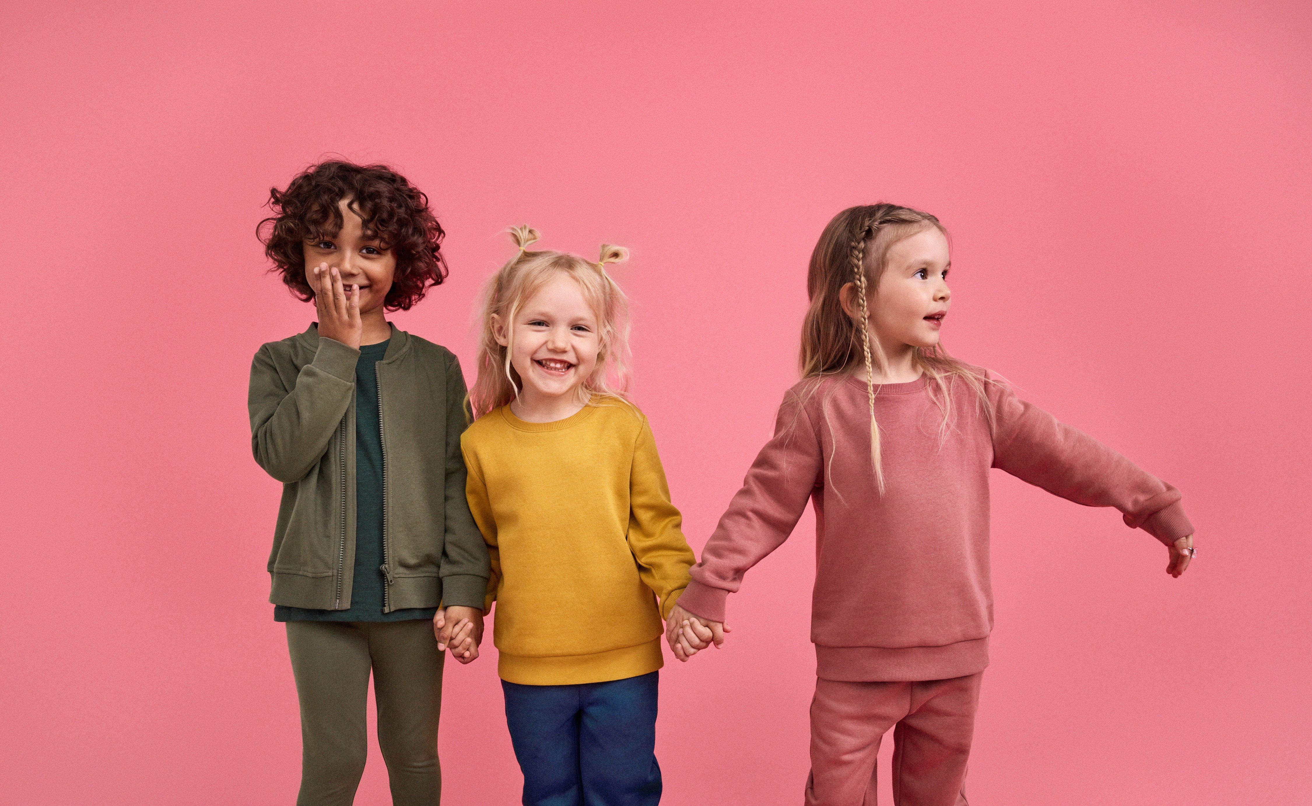  Three children stand in a row against a pink background. The first child is wearing pink clothes with his arms outstretched to the sides. The second child is wearing green clothes and making the same gesture. The third child is wearing blue pants and a yellow shirt, with one arm up and the other down.