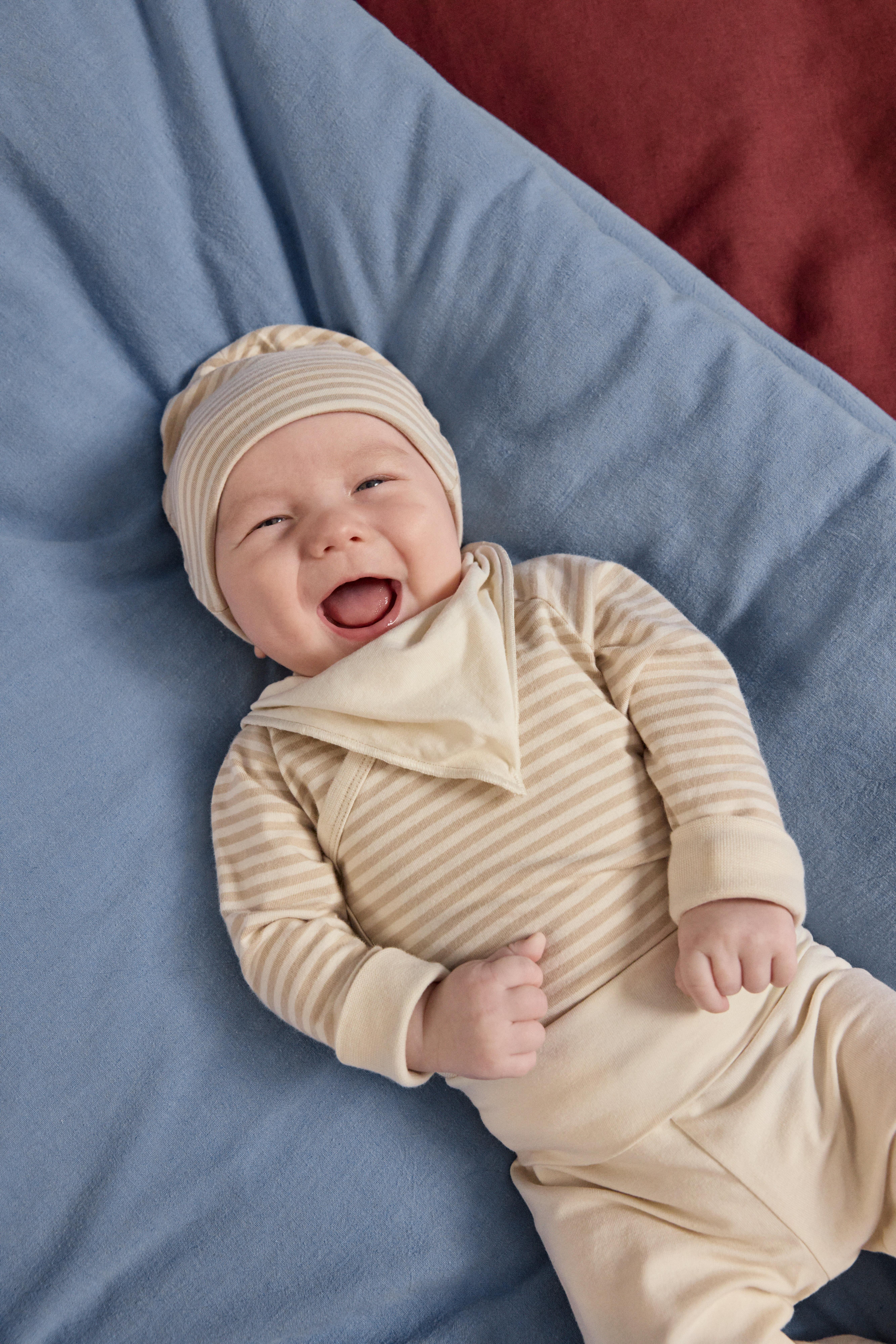 A smiling and happy baby lies on his back on a dark blue blanket. He is dressed in a soft and comfortable starter kit from Lindex.