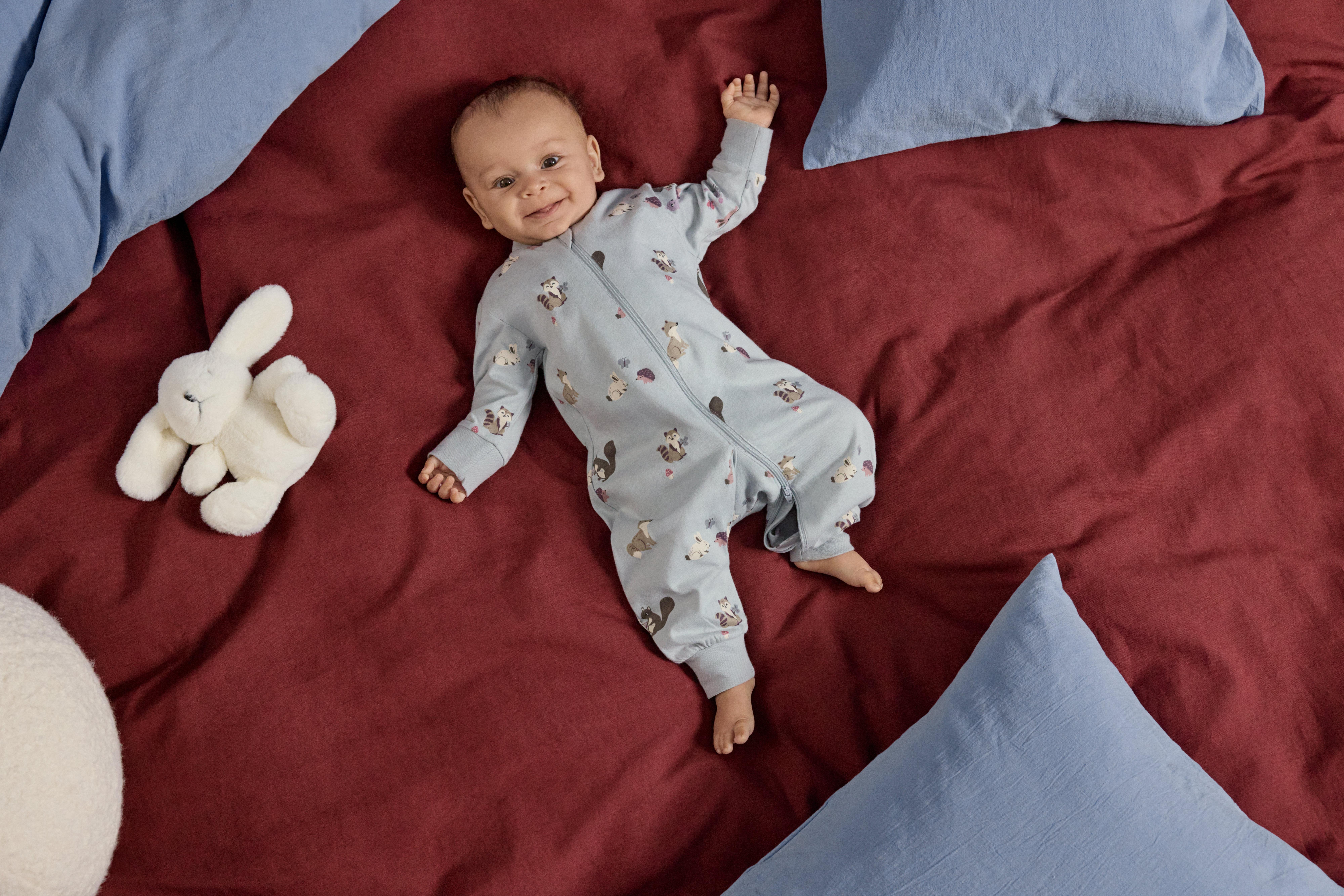A happy, laughing baby lies on his back on a red blanket. The baby is wearing light blue printed pajamas. Next to the baby is a white cuddly bunny.