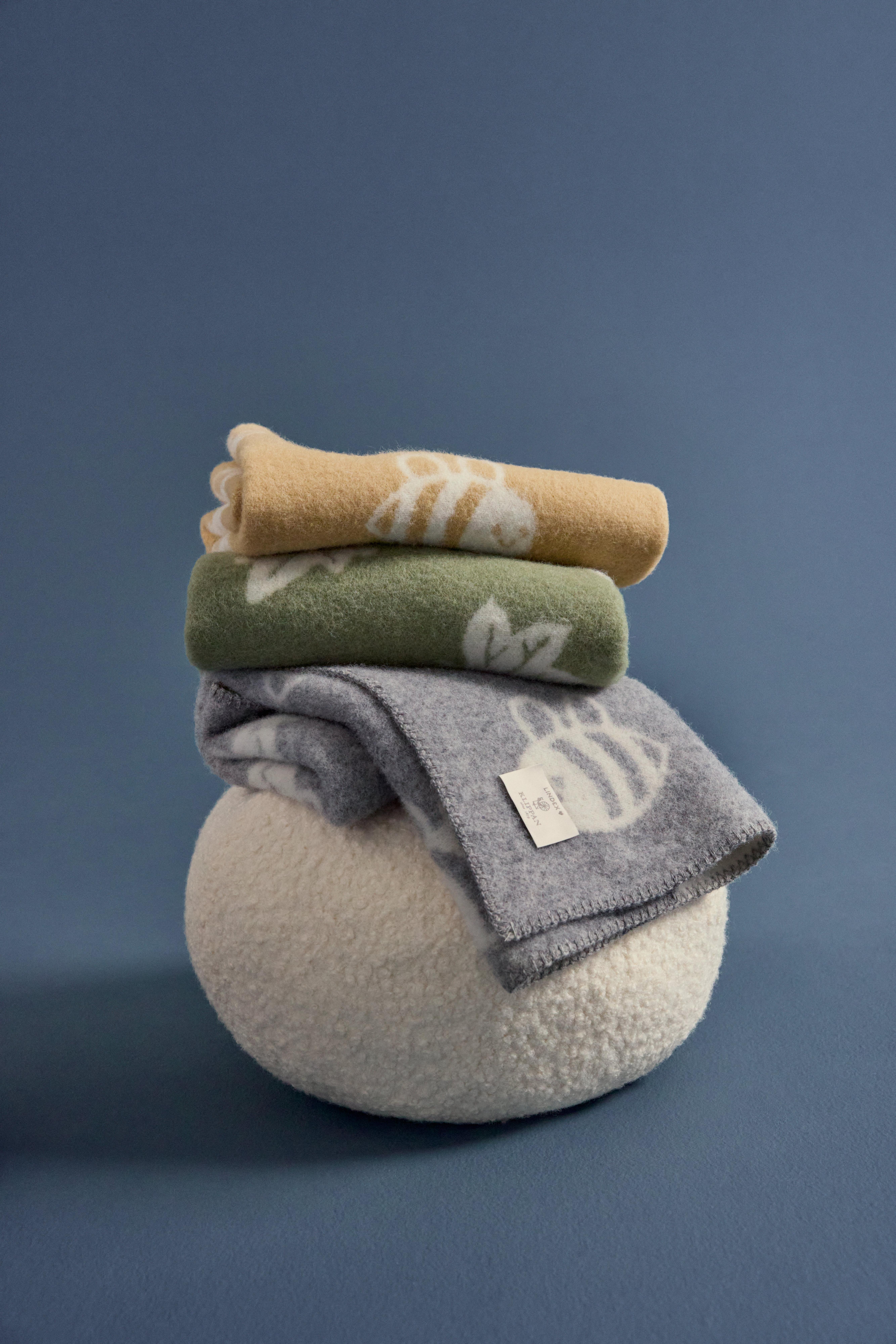  A stack of three baby blankets lies on a pouf. One is yellow with bees, one is green with leaves, and one is light gray with bees.