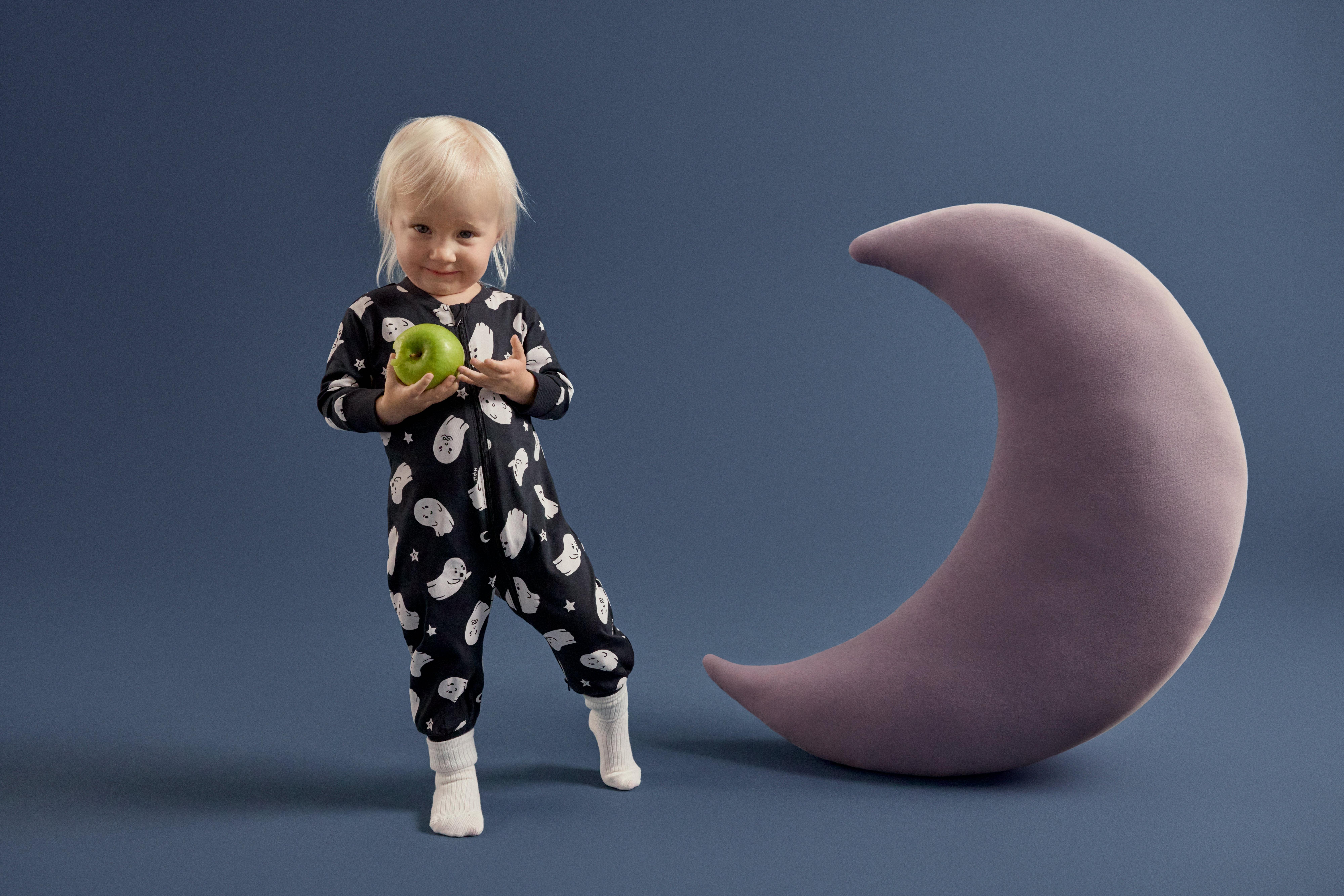 Malá holčička se plíží v pyžamu s potiskem duchů. Pozadí je tmavě modré a napravo je velká měkká hračka ve tvaru měsíce.