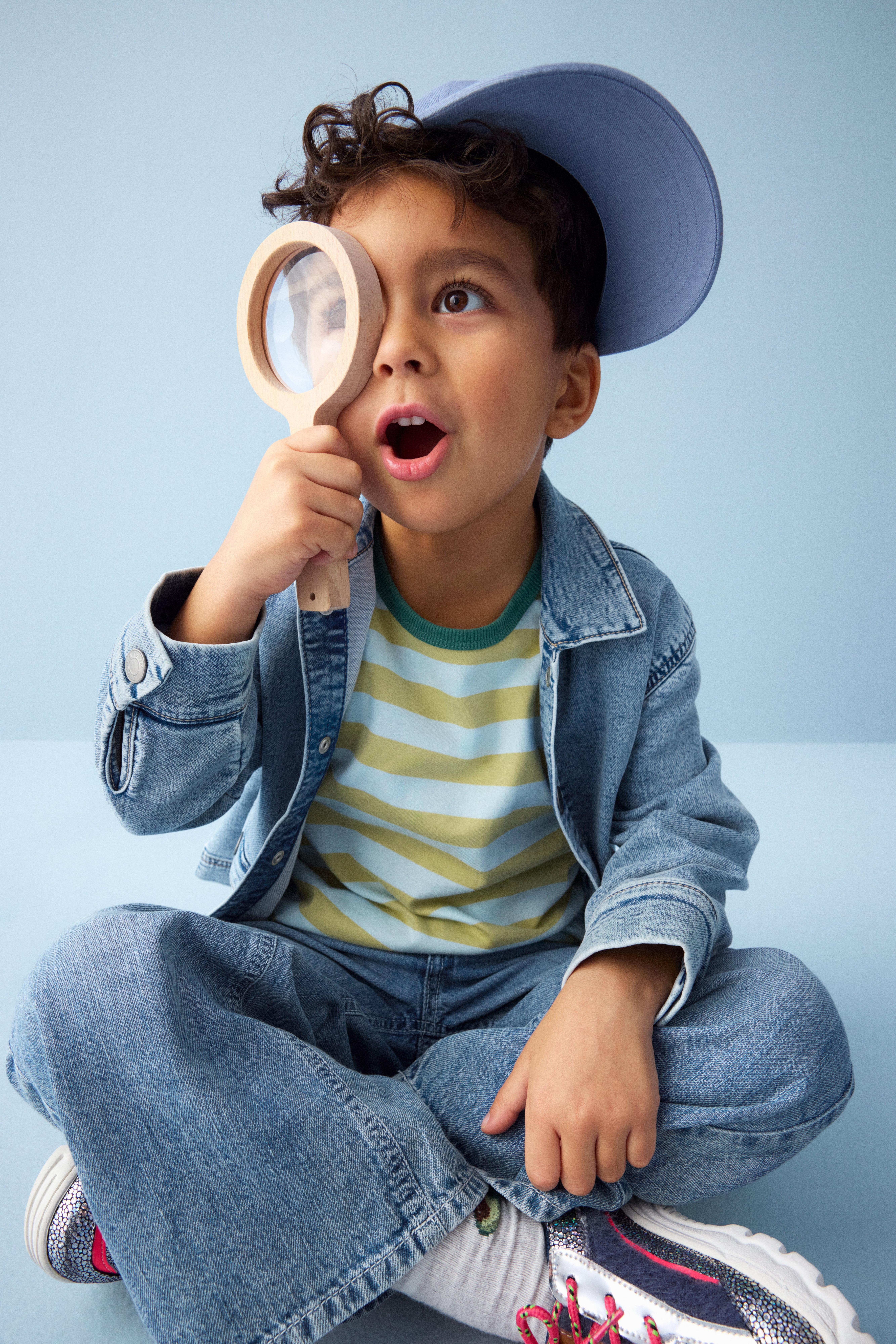 En bild på Albert, 5 år gammal, som bär en vit och gulrandig t-shirt, jeans, en jeansjacka och en blå keps. Han tittar i ett förstoringsglas.