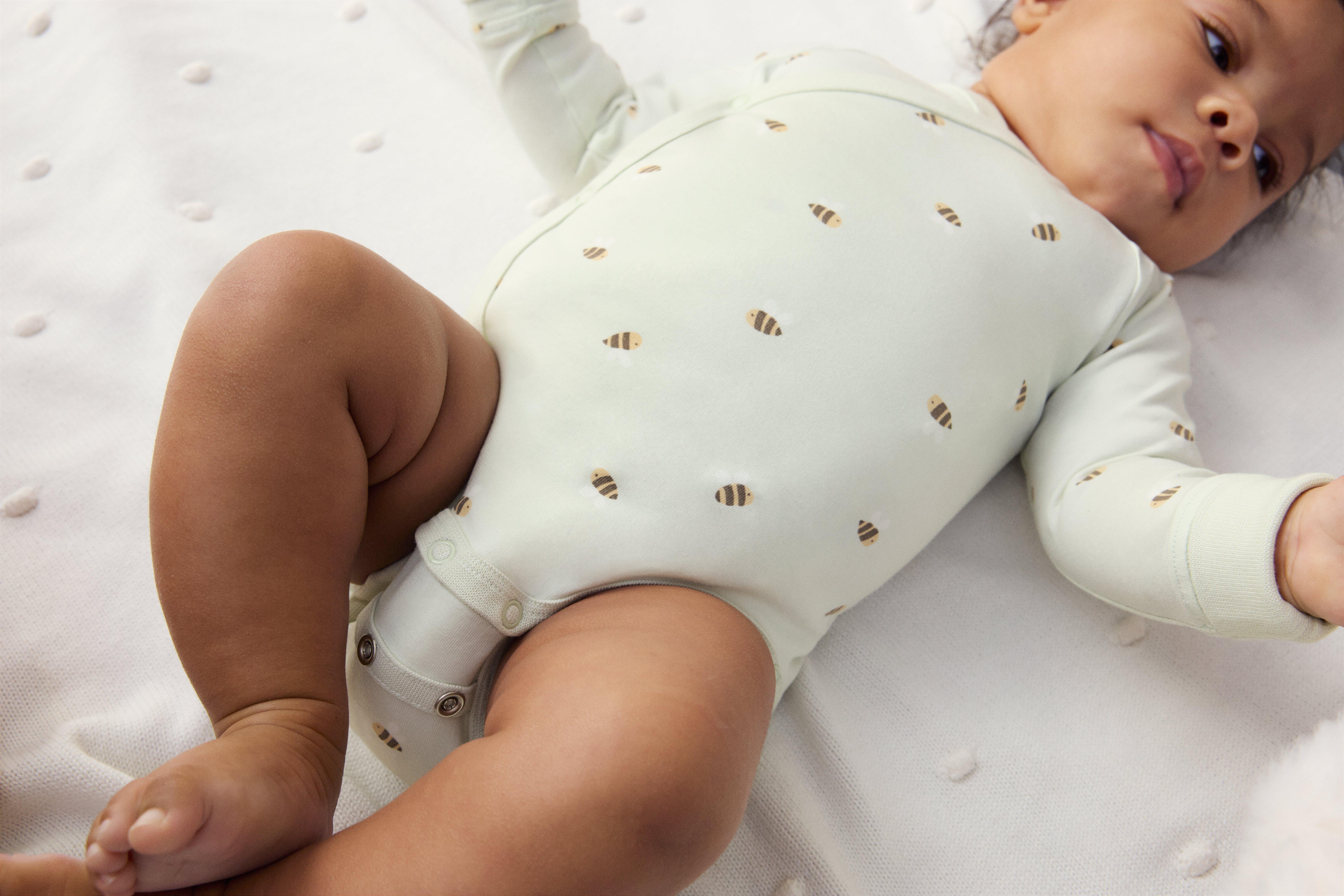 Bábätko oblečené v béžovom body s potlačou malých chlpatých oviec. Bábätko leží na tmavočervenej deke.