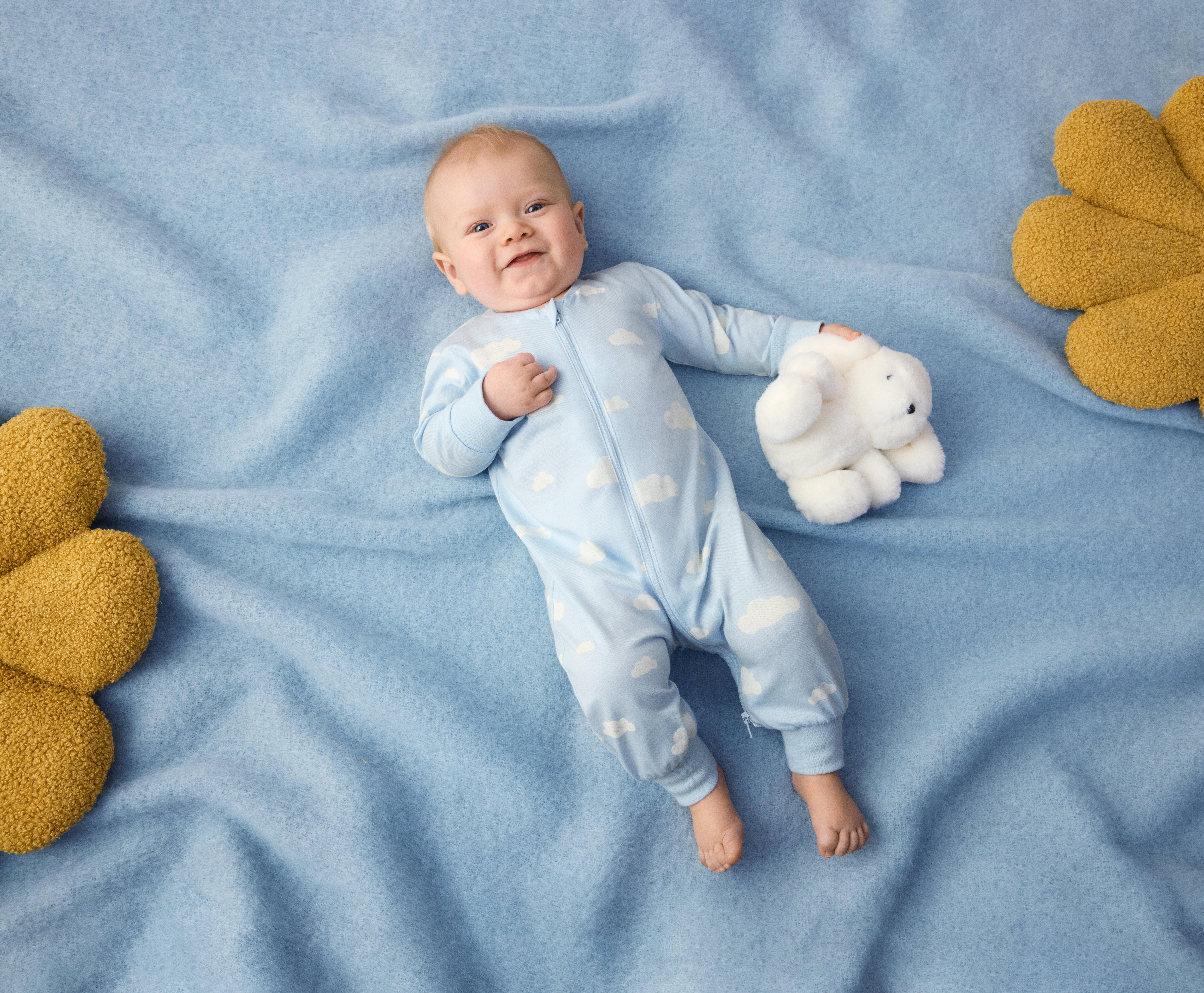 A happy, laughing baby lies on his back on a light blue blanket. The baby is wearing light blue printed pajamas. Next to the baby is a white cuddly bunny.