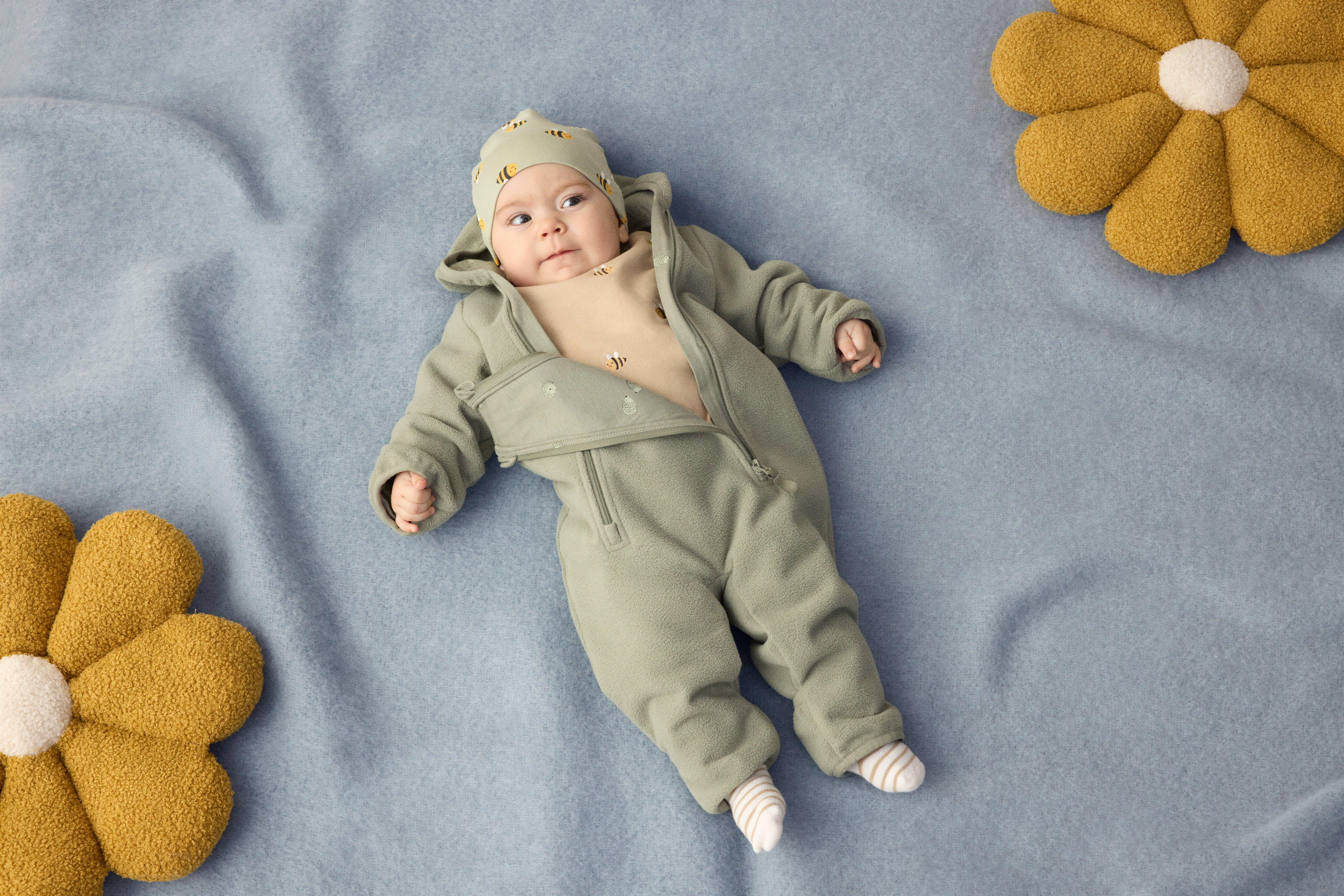 En baby ligger på en blå filt. Babyn är klädd i en öppnad overall och en mössa på huvudet.