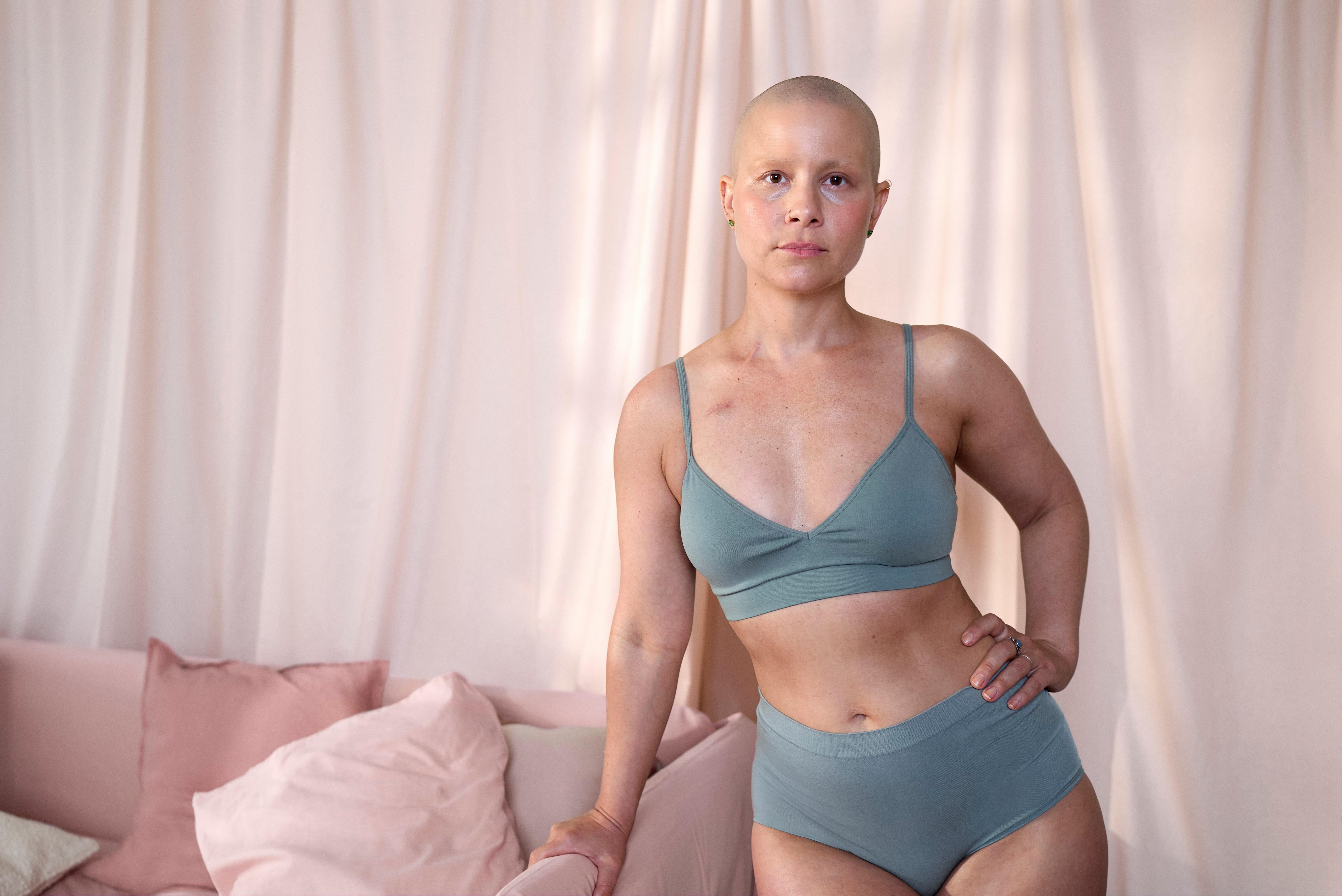  A woman, dressed in Dusty Green colored underwear, stands leaning against a pink sofa. Behind her is a pink fabric background wall. The woman has no hair and looks seriously straight into the camera.