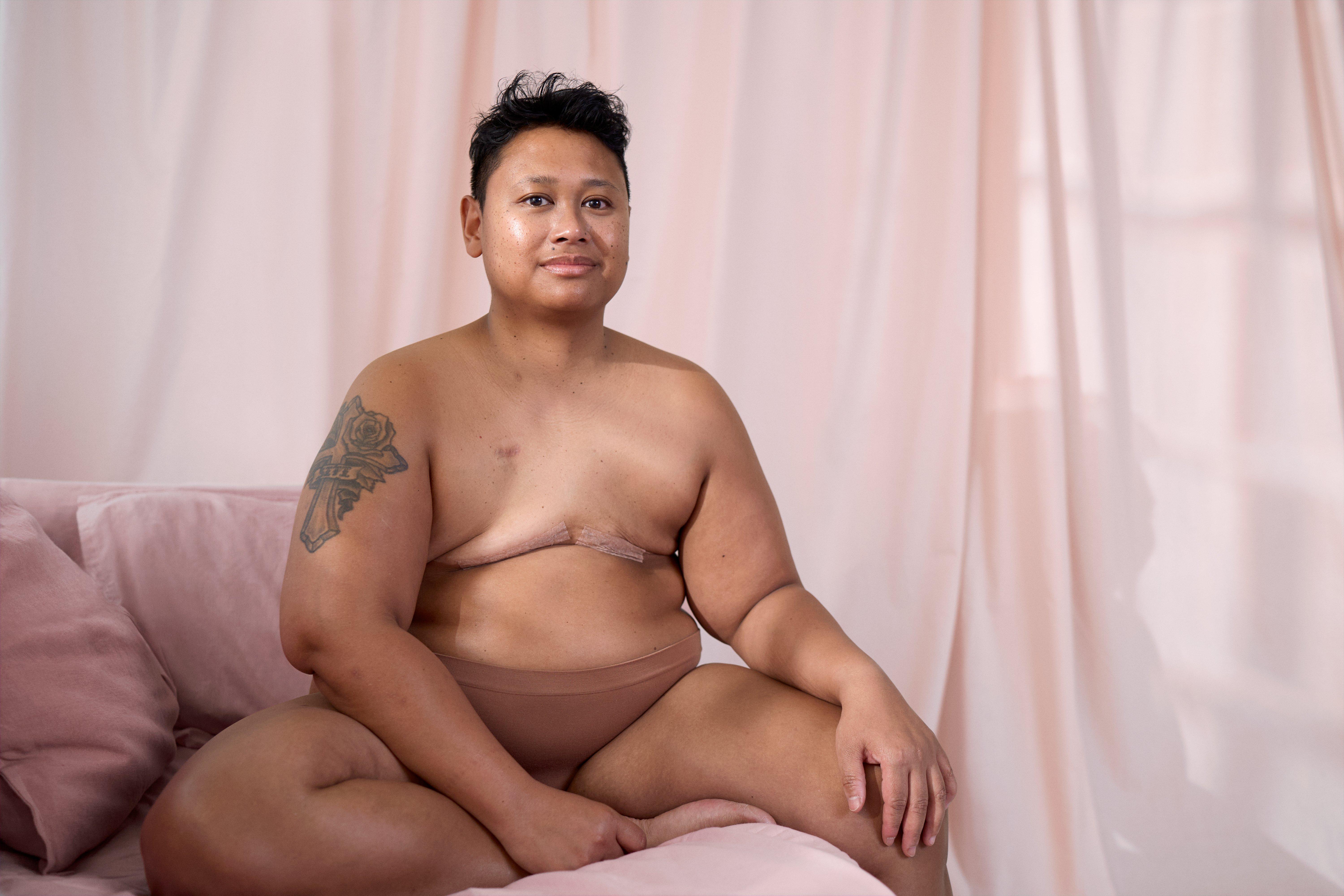  A woman who has had a mastectomy sits curled up on a pink couch. She looks into the camera and gives a small smile.