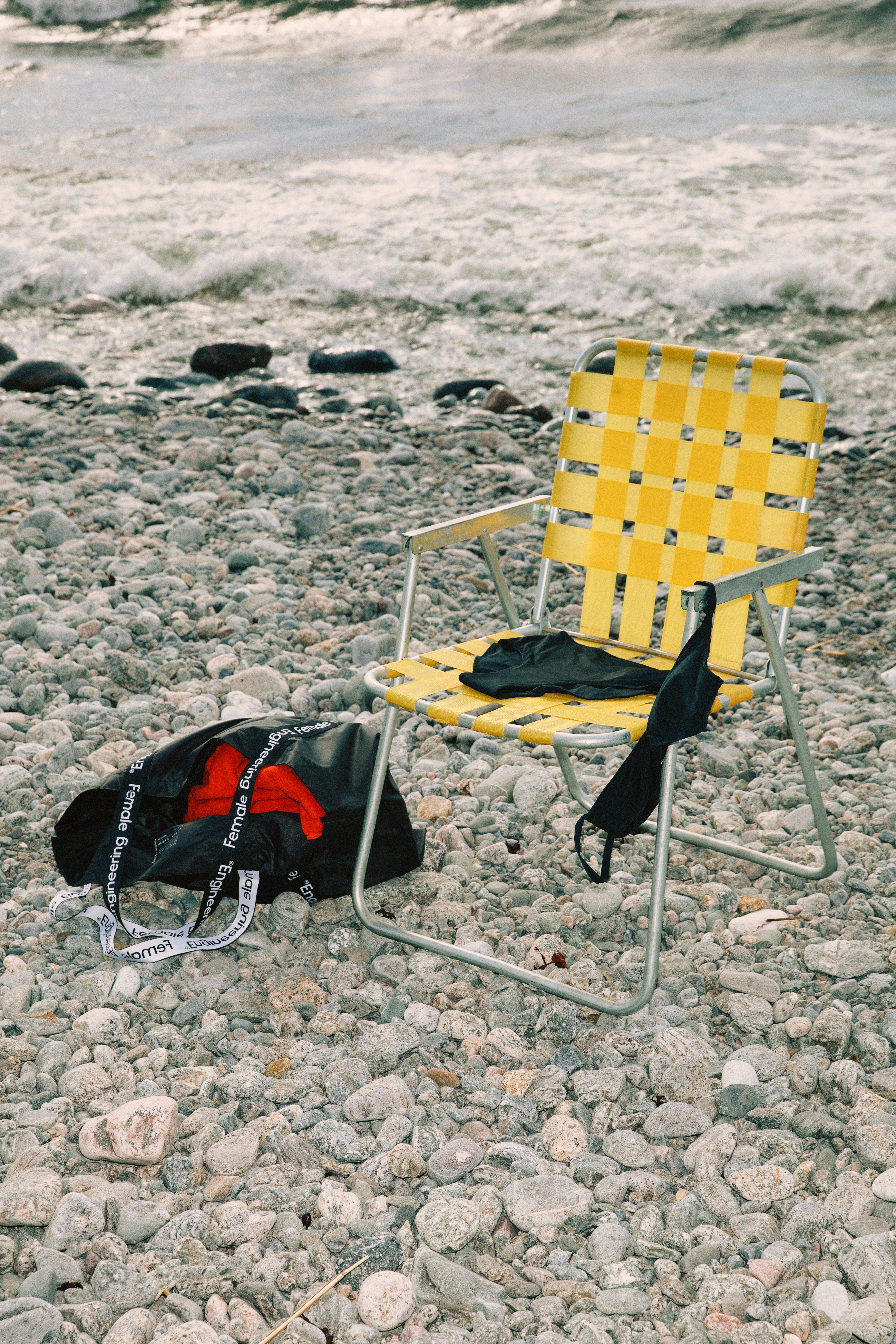 A yellow deck chair stands on a rocky beach. A black bikini is drying on the deck chair. On the ground is an open bag with a red towel.
