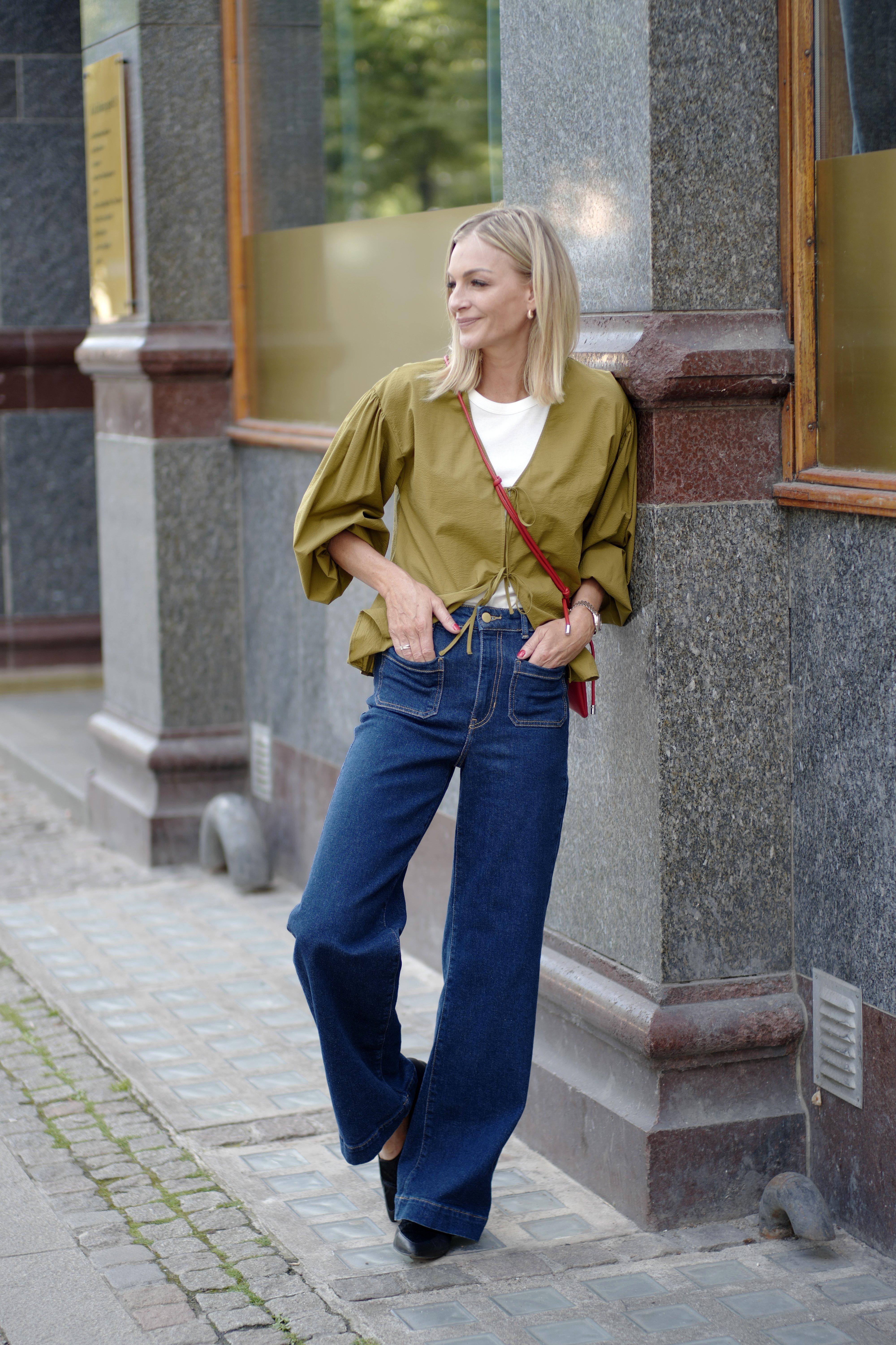 Kathrine, standing casually leaning against a stone wall, is wearing a green blouse with tie details, blue jeans and a white top.