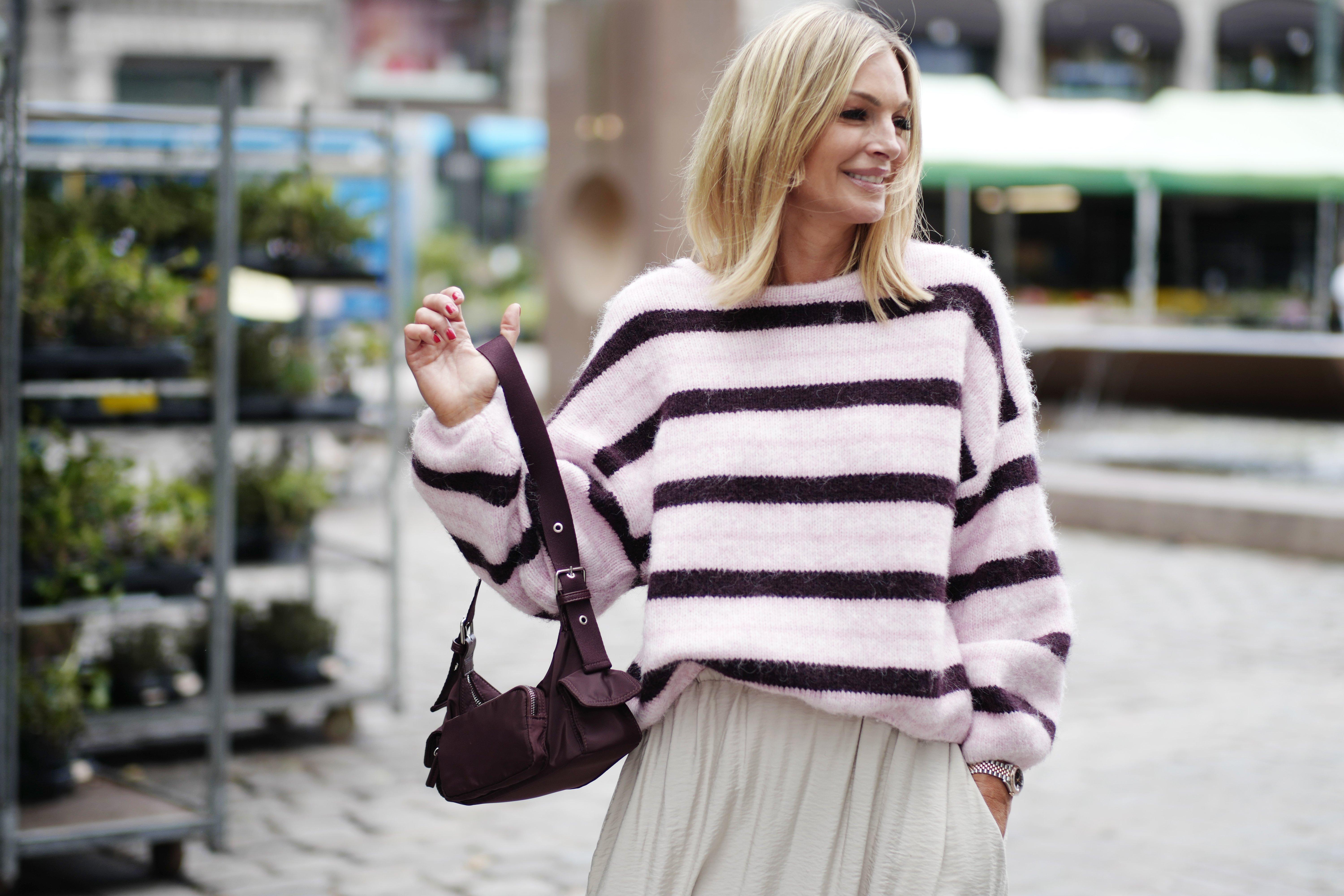  Kathrine, dressed in a cozy pink striped sweater and a long beige skirt. She is holding a brown shoulder bag in her hand.
