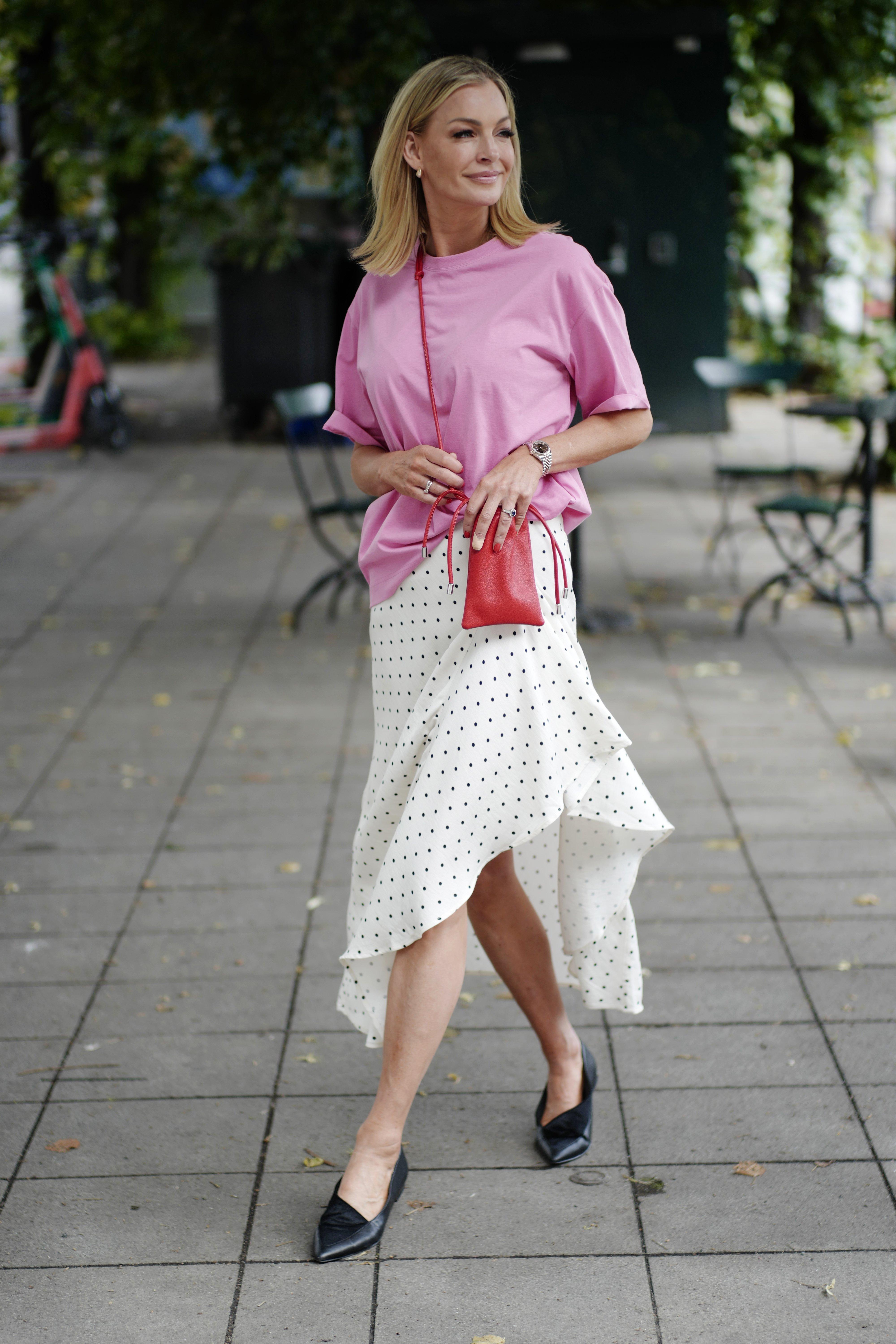 Kathrine, dressed in a white polka-dot skirt and pink t-shirt, walks across an outdoor café. She has a red mobile phone bag over her shoulder.