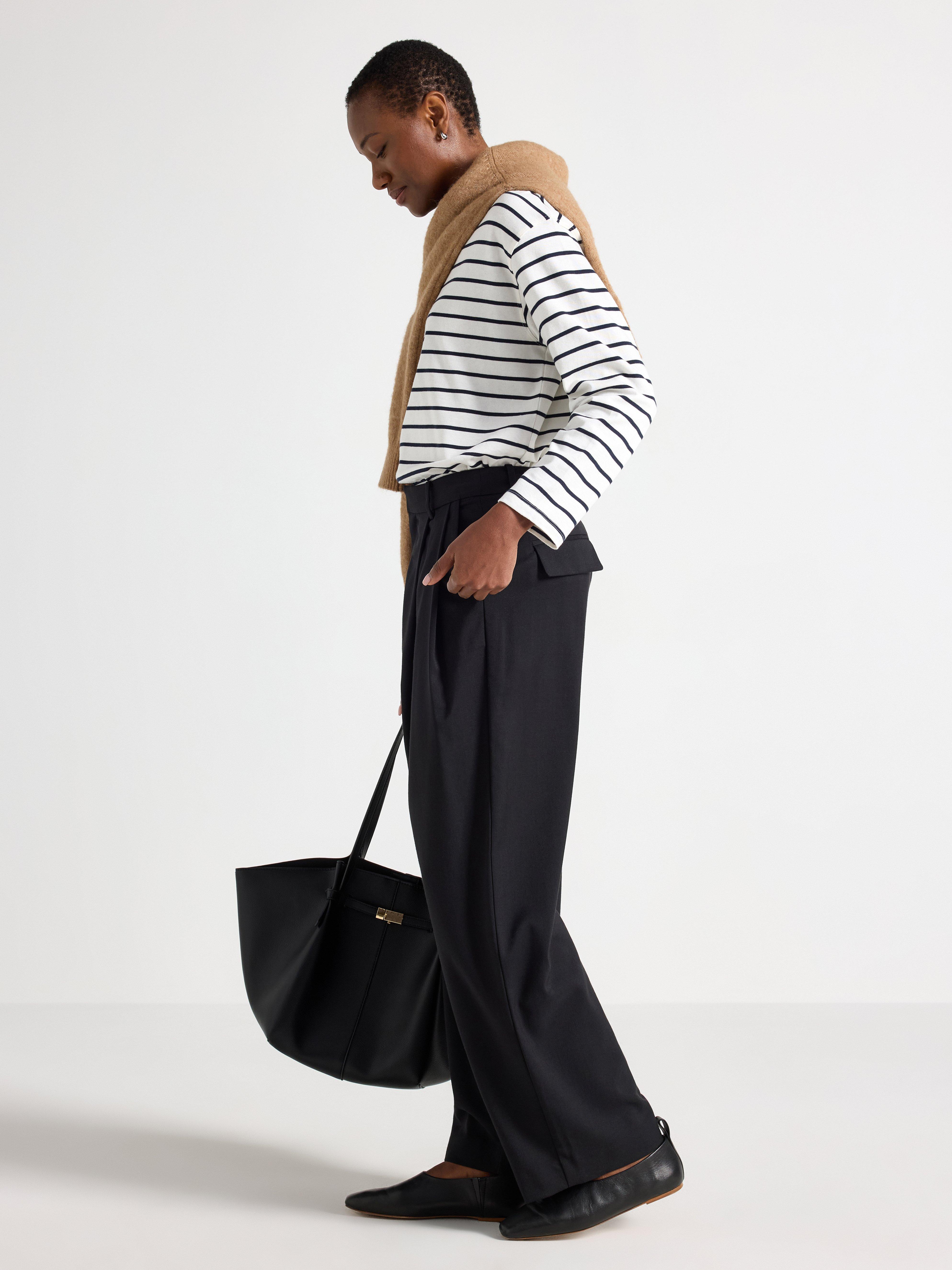 A woman photographed from the side. Dressed in a white and black striped top, black pants and with a beige sweater over her shoulders, she is holding a bag in her hand.